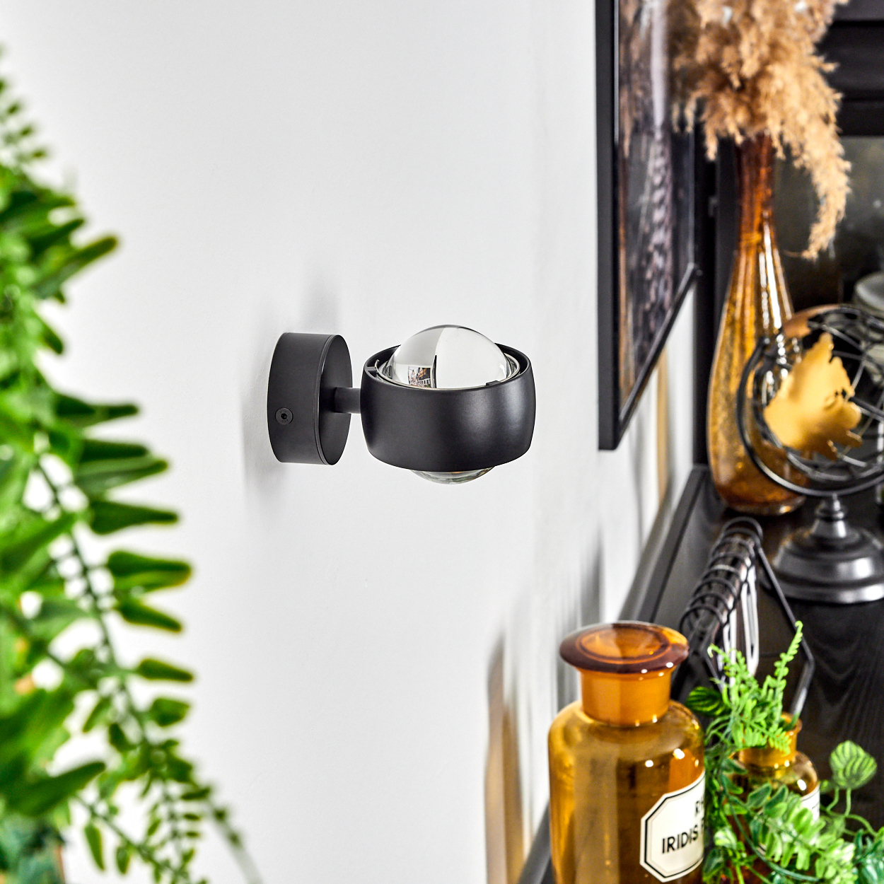 Eine schwarze Wandlampe mit kugelförmiger Glühbirne an weißer Wand. Weitere Dekoartikel sind im Hintergrund zu sehen.