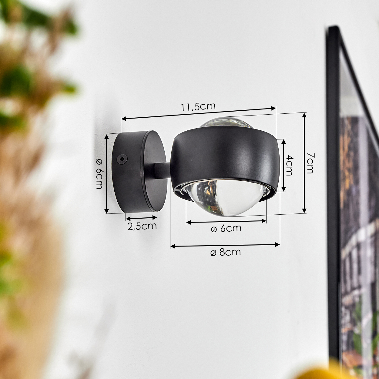 Eine schwarze Wandleuchte mit kugelförmigem Glasschirm ist an einer weißen Wand befestigt. Abmessungen sind angegeben. Rechts ist ein gerahmtes Bild.