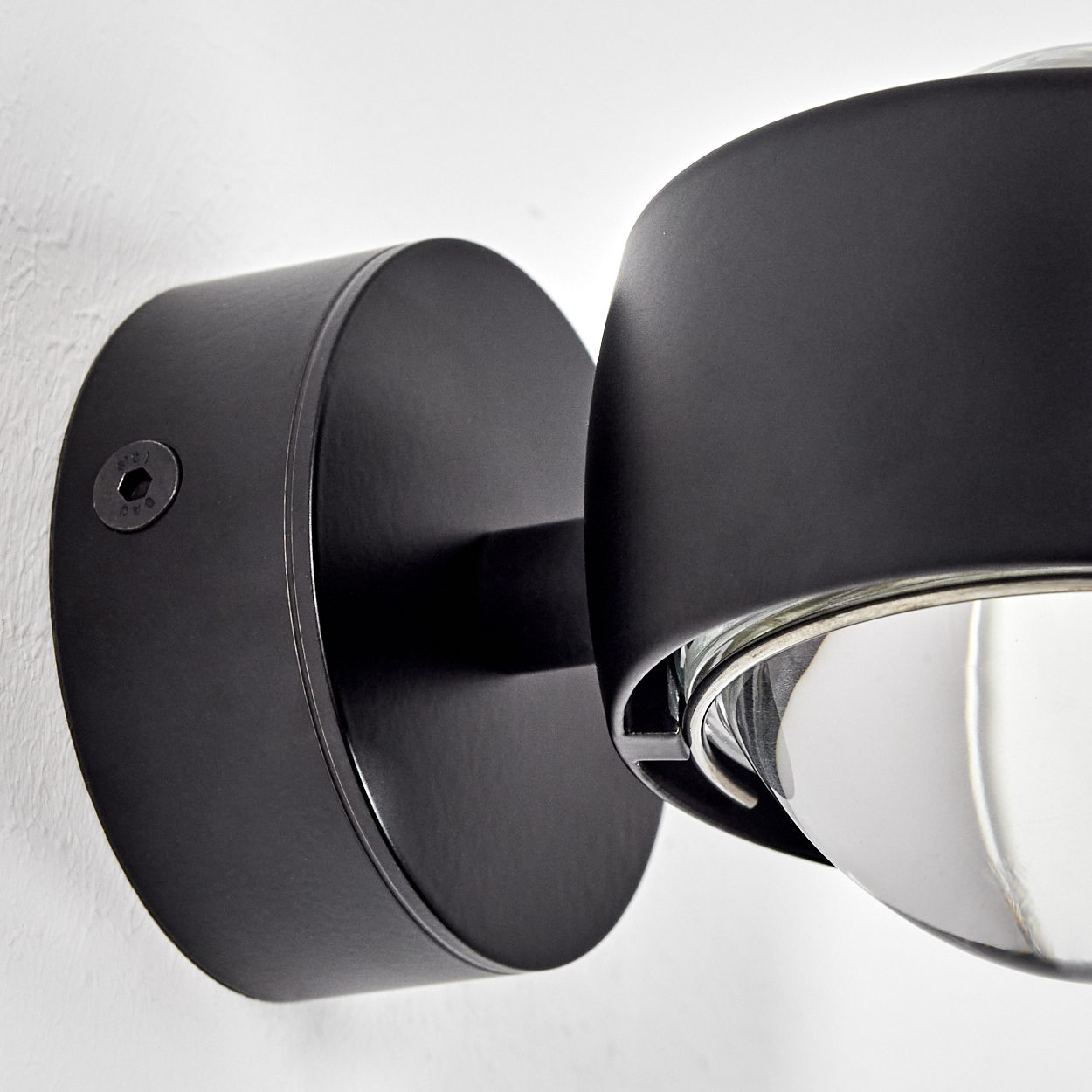Nahaufnahme einer schwarzen Wandleuchte mit einer Glaskugel an einer weißen Wand. Die Lampe hat einen runden Sockel und einen geschwungenen Schirm.