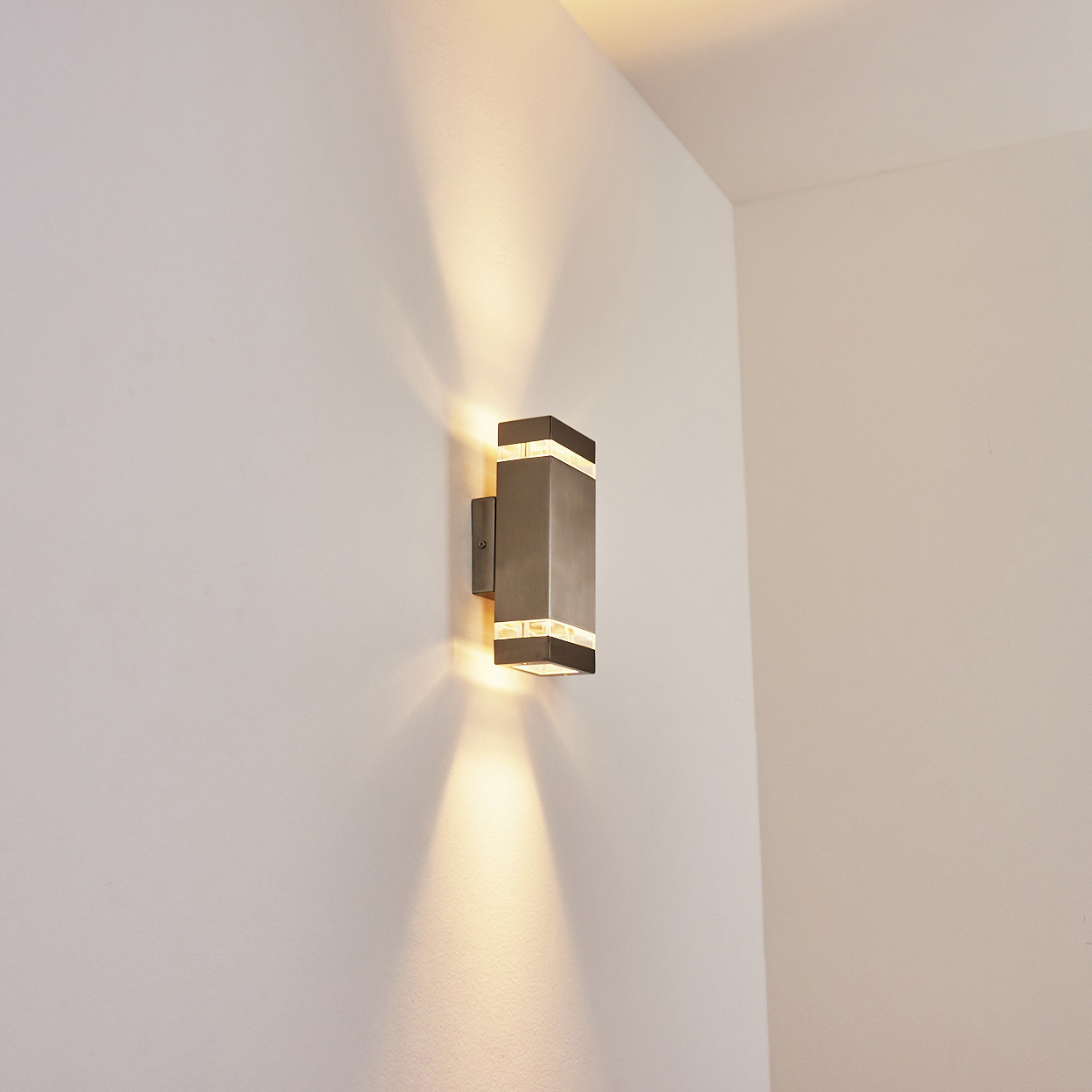 Eine moderne, rechteckige Wandlampe mit warmem Licht ist an einer weißen Wand montiert. Die Lampe strahlt Licht nach oben und unten aus.