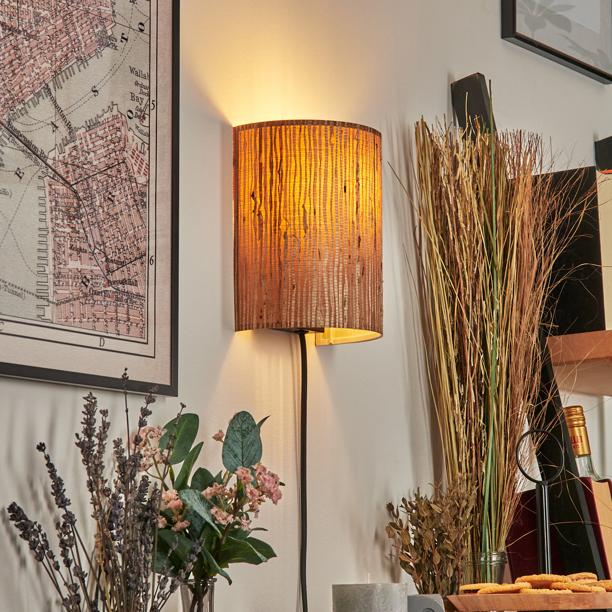 Eine beleuchtete Wandlampe, Karte, Blumen und Dekorationen. Der Lampenschirm ist braun und strukturiert. Blumen und eine Karte hängen an der Wand.