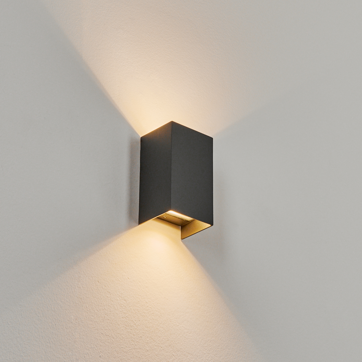 Eine quadratische, dunkelgraue Wandleuchte an einer weißen Wand. Die Lampe strahlt warmes Licht aus, das Schatten wirft.