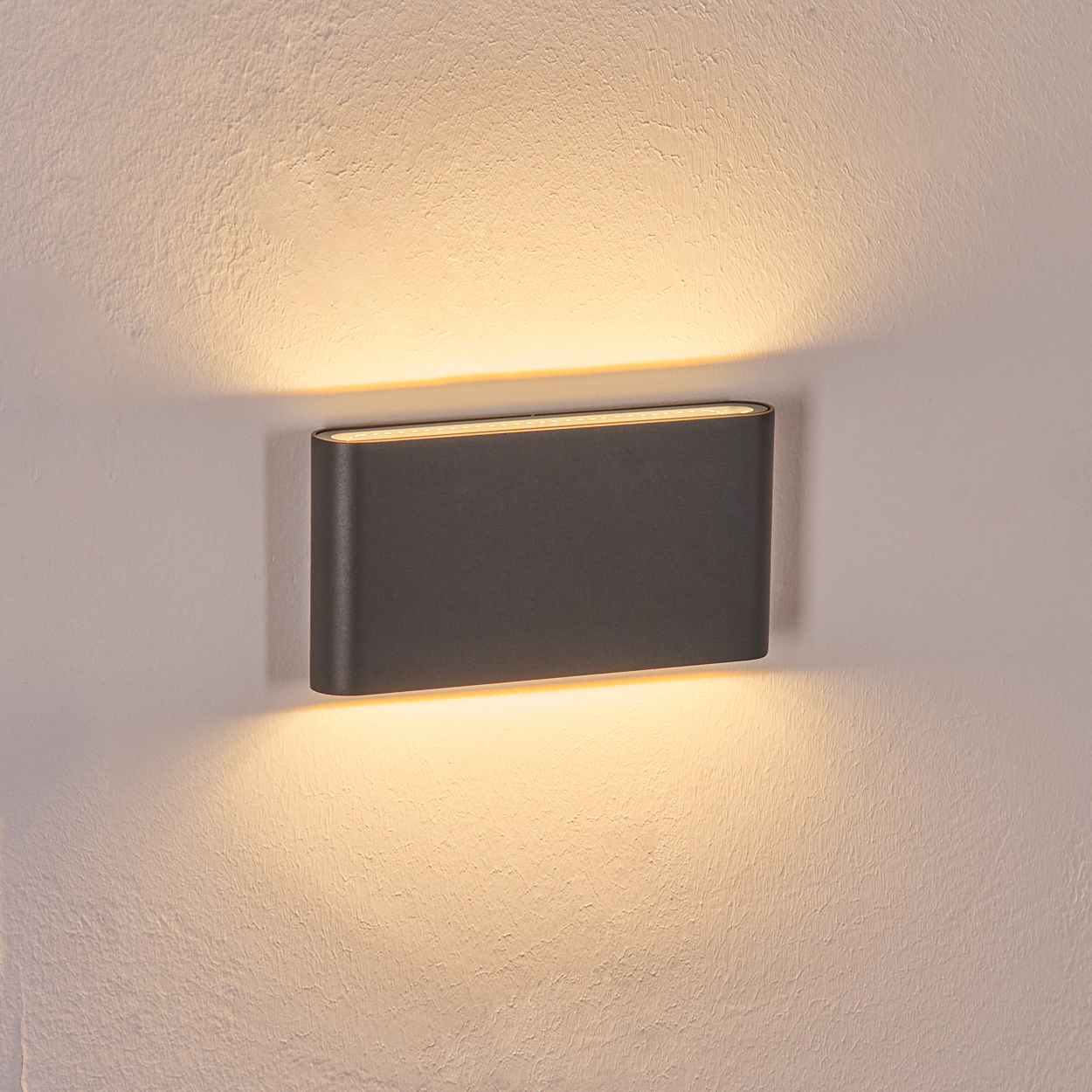 Eine dunkelgraue rechteckige Wandlampe ist an einer strukturierten weißen Wand befestigt. Die Lampe strahlt warmes Licht aus.
