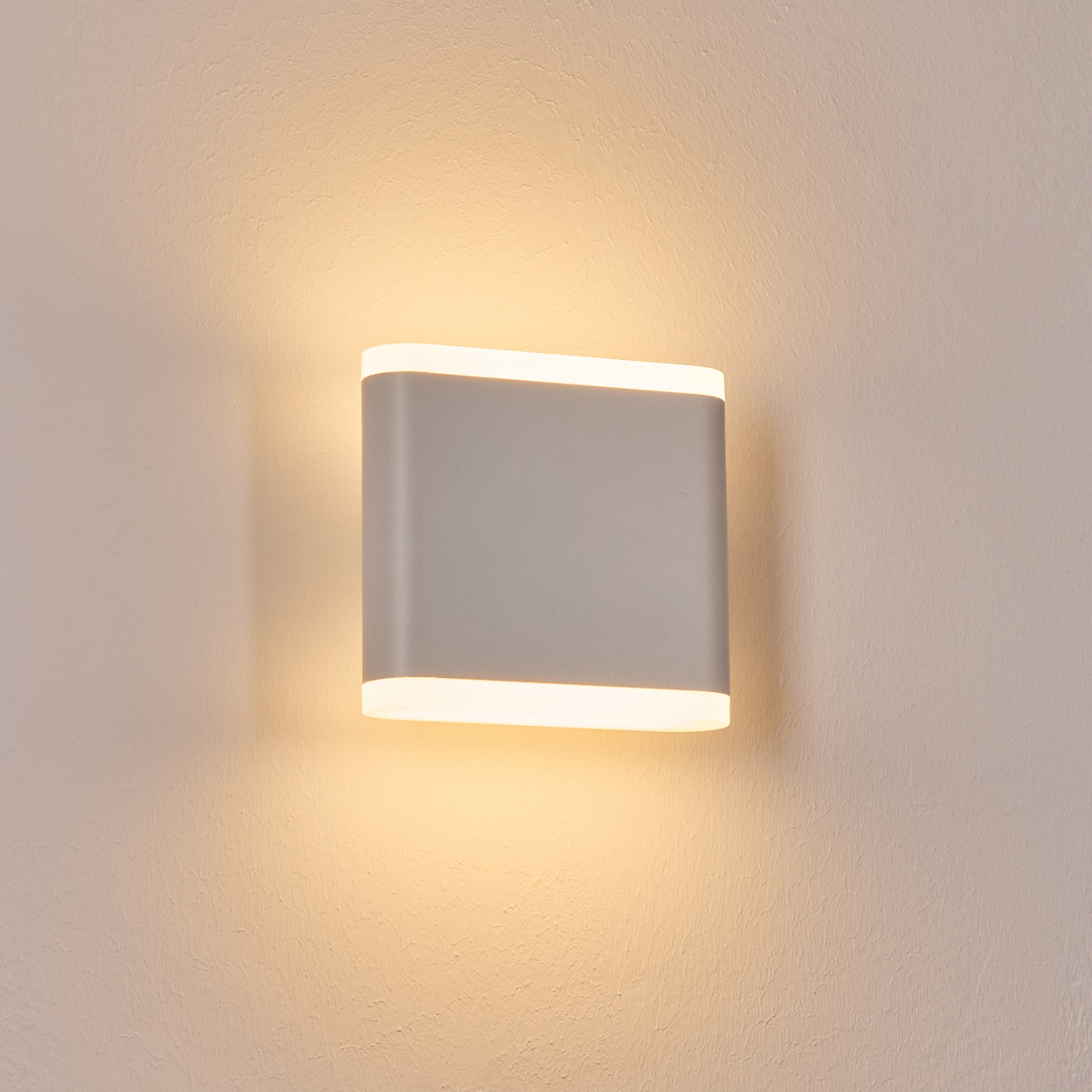 Eine moderne, rechteckige Wandleuchte mit weichem, diffusem Licht, montiert an einer strukturierten, cremefarbenen Wand. Die Lampe strahlt warm.