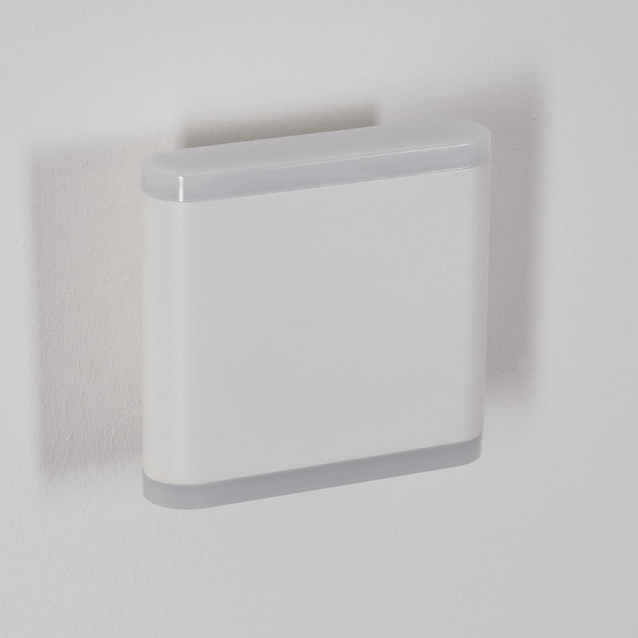 Eine rechteckige weiße Wandleuchte mit grauem Rand ist an einer weißen Wand montiert. Die Lampe hat eine abgerundete Ober- und Unterseite.