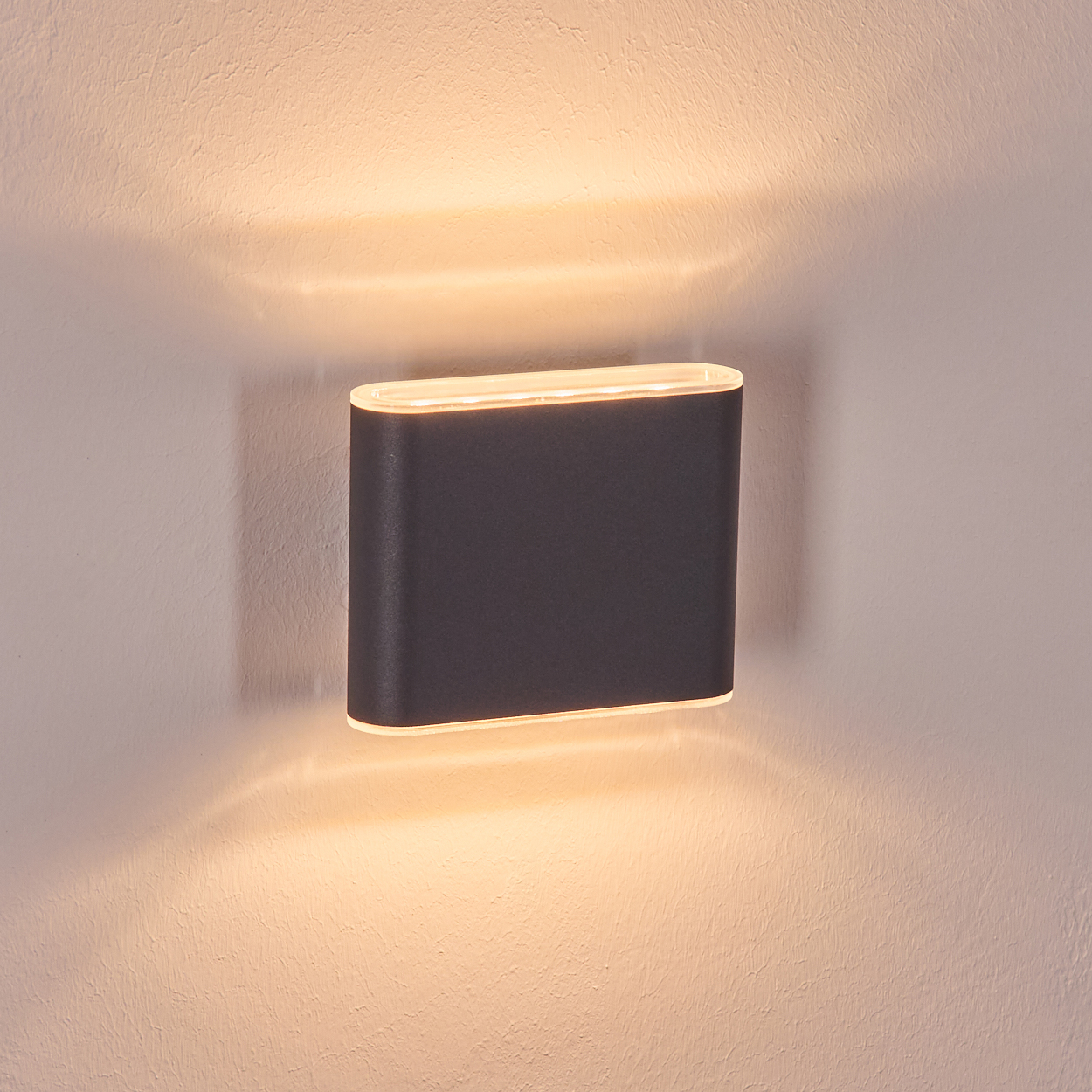 Eine dunkelgraue Wandleuchte mit rechteckiger Form ist an einer hellbeige Wand angebracht. Die Lampe strahlt warmes Licht aus.
