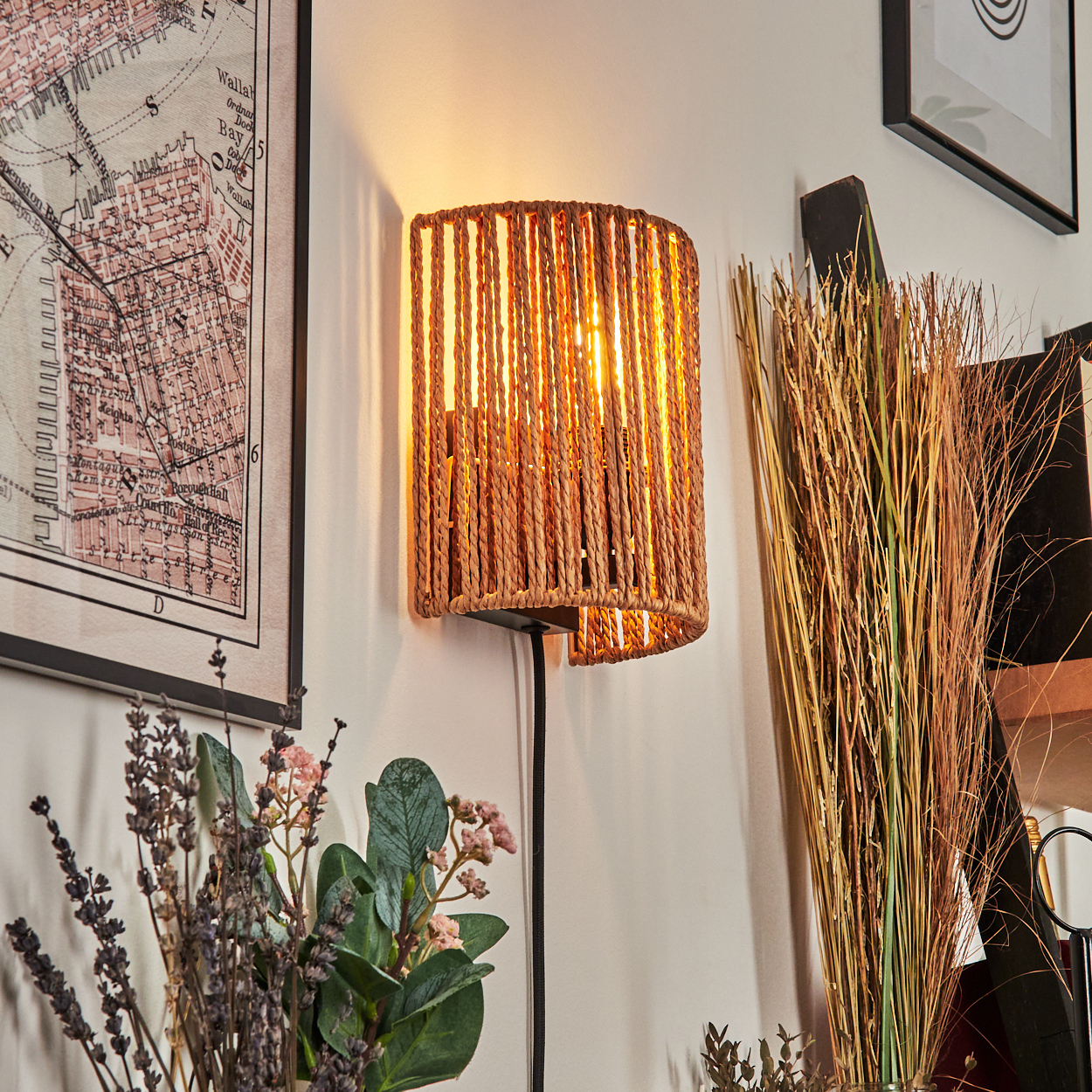 Eine gewebte Lampe beleuchtet eine Wand. Eine Karte und gerahmte Kunstwerke befinden sich neben der Lampe. Getrocknete Pflanzen und Blumen sind im Vordergrund.