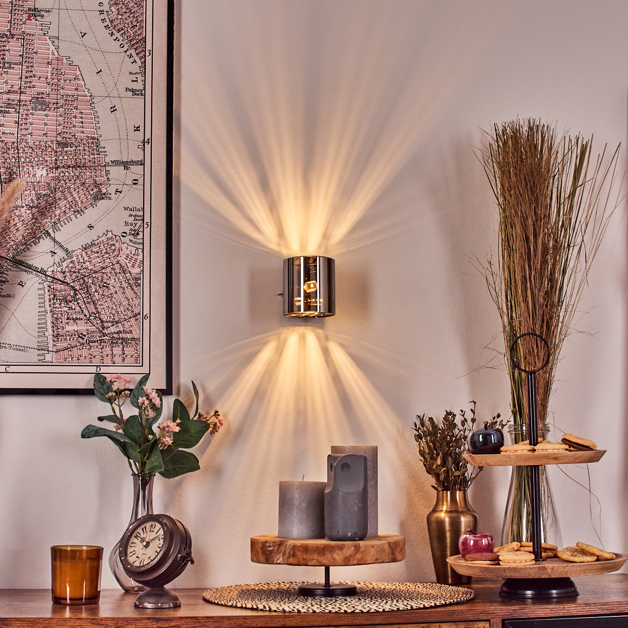 Eine Wandlampe scheint Licht auf eine weiße Wand. Auf einem Tisch befinden sich eine Karte, eine Uhr, Kerzen, Blumen und ein Tablett mit Keksen.
