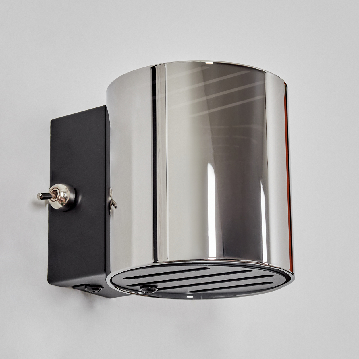 Eine moderne, verchromte Wandlampe mit zylindrischem Schirm und schwarzem, rechteckigem Sockel. Auf der linken Seite ist ein Schalter zu sehen.