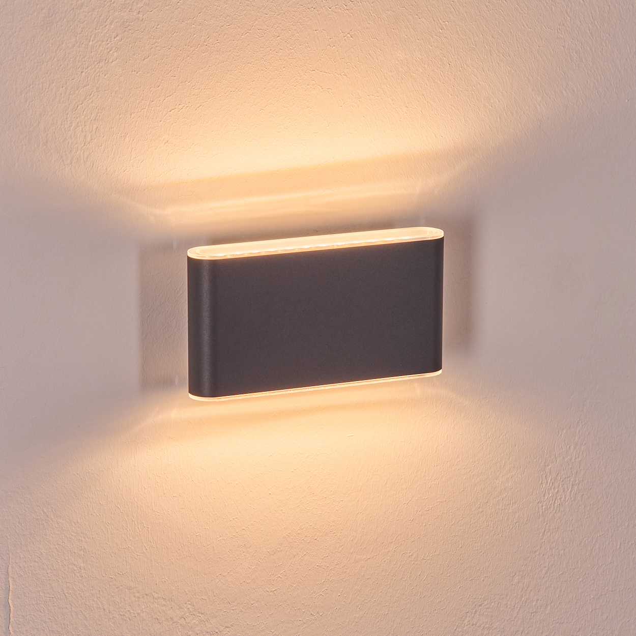 Eine dunkelgraue, rechteckige Wandleuchte ist an einer strukturierten weißen Wand angebracht. Die Lampe strahlt helles Licht aus.