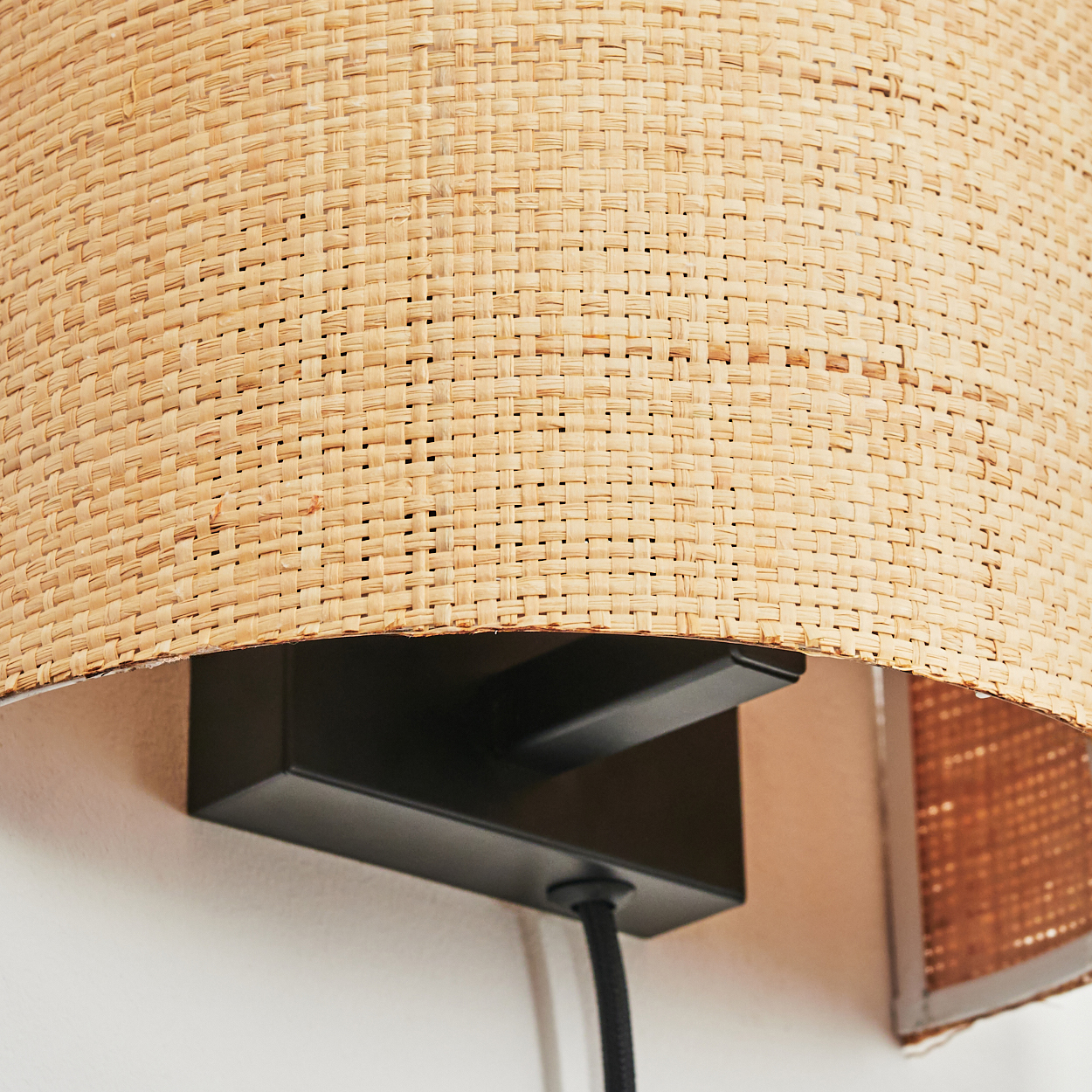 Ein Lampenschirm aus gewebtem Rattan ist auf einem schwarzen Metallsockel montiert. Ein schwarzes Kabel hängt darunter. Der Hintergrund ist eine weiße Wand.