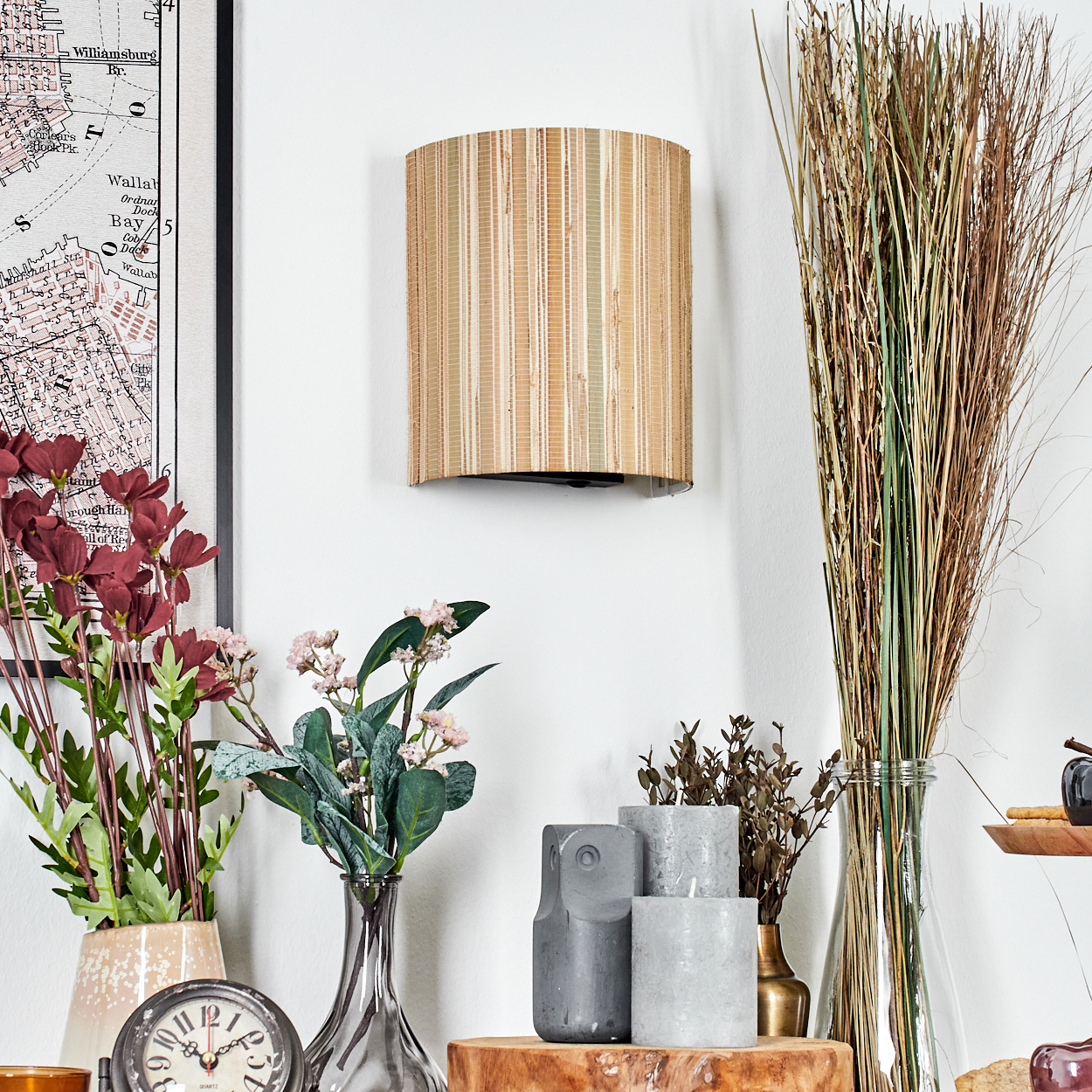 Eine Wandlampe aus Bambus, Blumen in Vasen, Kerzen, eine Karte und hohes Gras in einer Glasvase sind vor einer weißen Wand angeordnet.