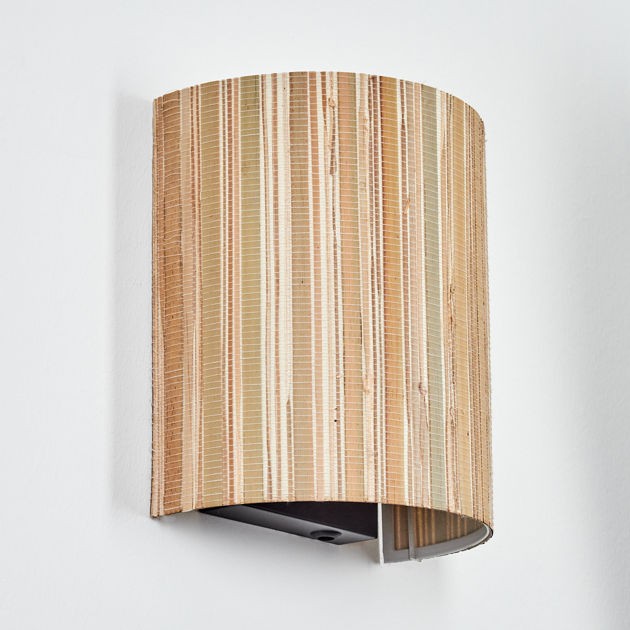 Eine Wandleuchte mit gewebtem, zylindrischem Schirm. Der Schirm ist braun und beige. Sie ist an einer weißen Wand befestigt.