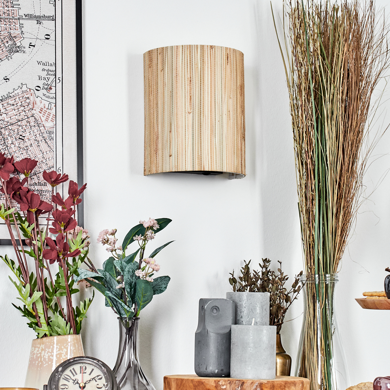 Eine Wandlampe, Blumen und eine Karte an einer weißen Wand. Darunter Vasen, eine Uhr, Kerzen und hohes Gras in einer Vase.