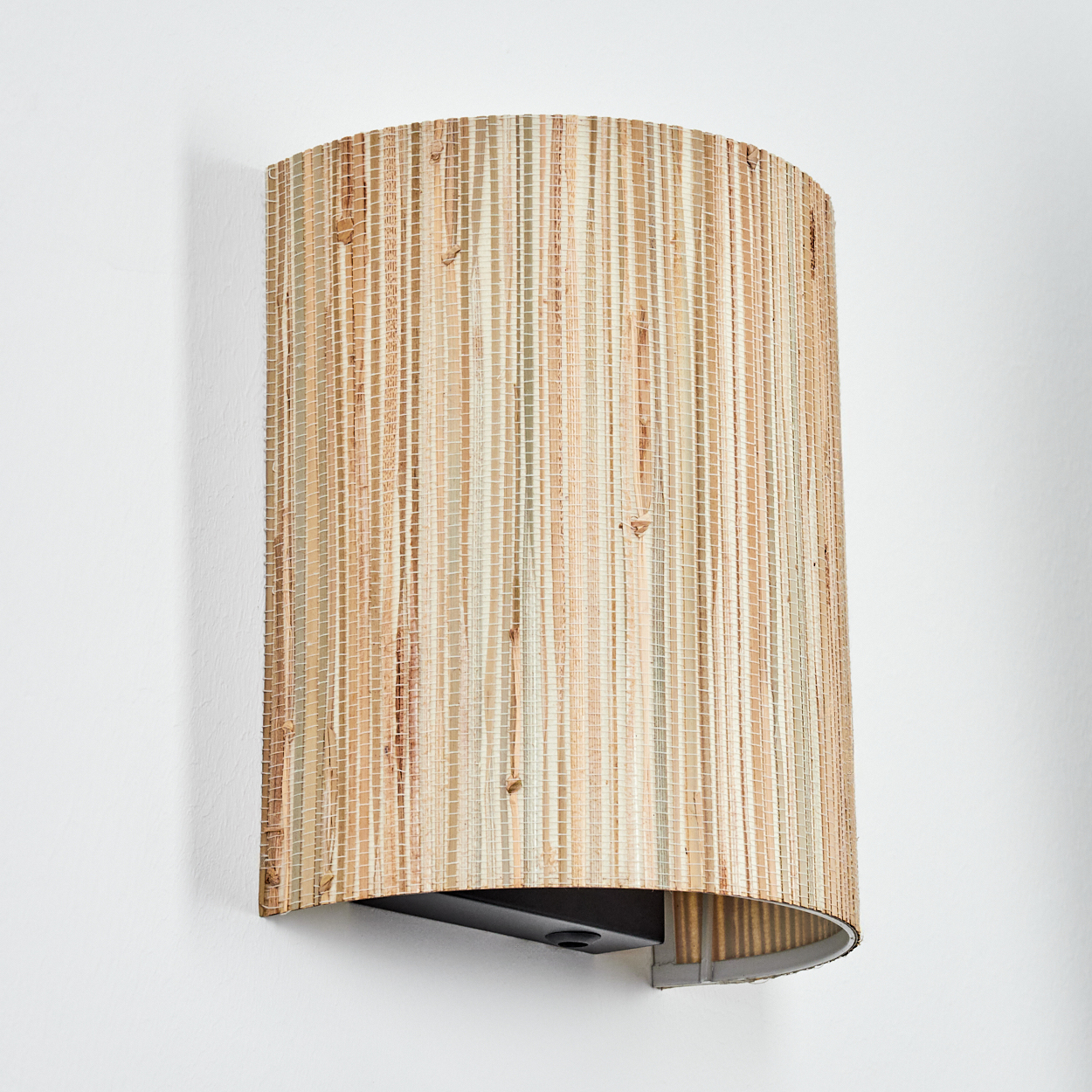 Eine Wandleuchte mit einem zylindrischen Lampenschirm aus gewebten beigen und cremefarbenen Naturfasern, an einer weißen Wand.