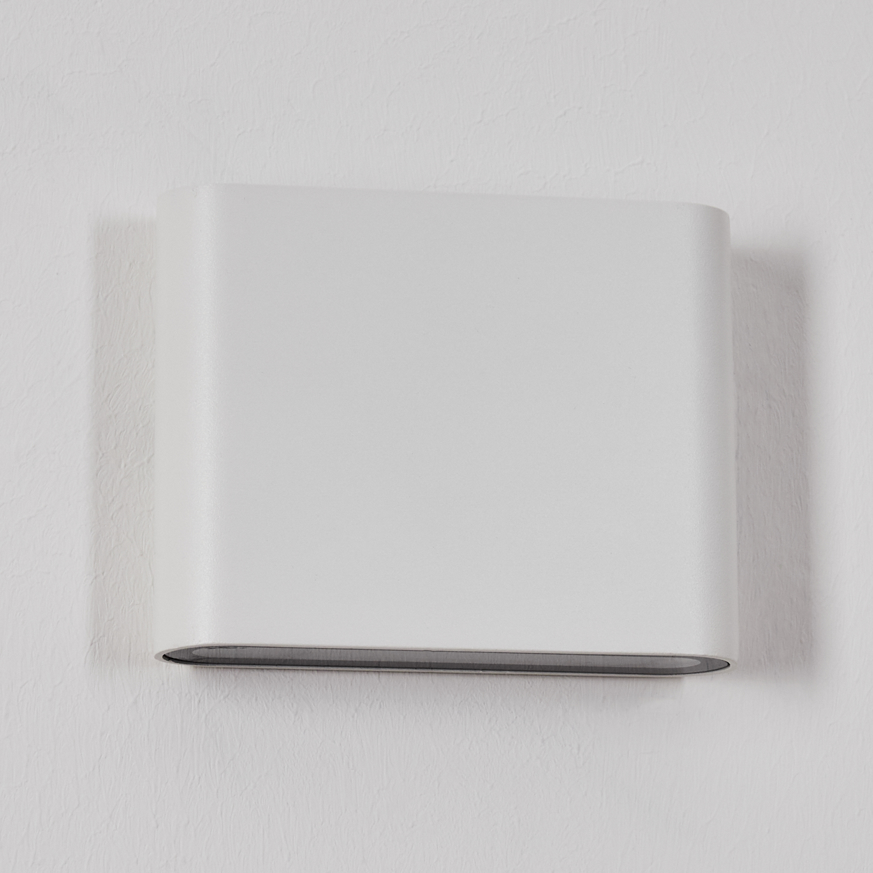 Eine weiße, rechteckige Wandleuchte ist an einer strukturierten weißen Wand befestigt. Die Lampe hat eine gewölbte Ober- und Unterseite mit grauem Akzent.