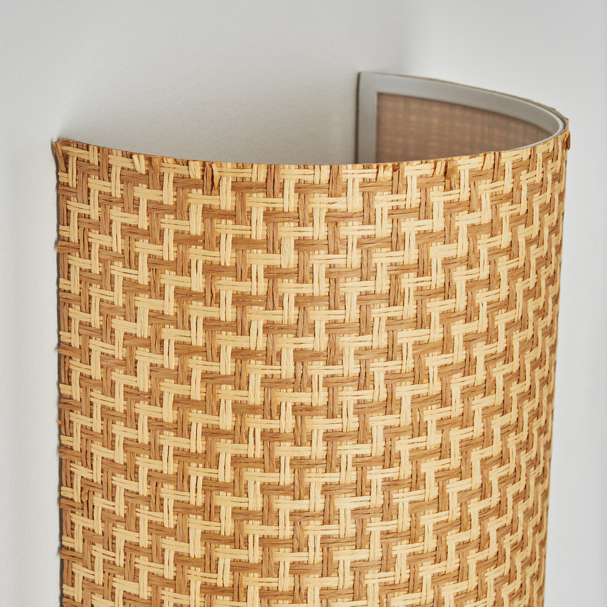 Eine gewebte, halb zylindrische Wandleuchte. Die Lampe besteht aus tan- und braun gewebtem Material und ist an einer weißen Wand befestigt.