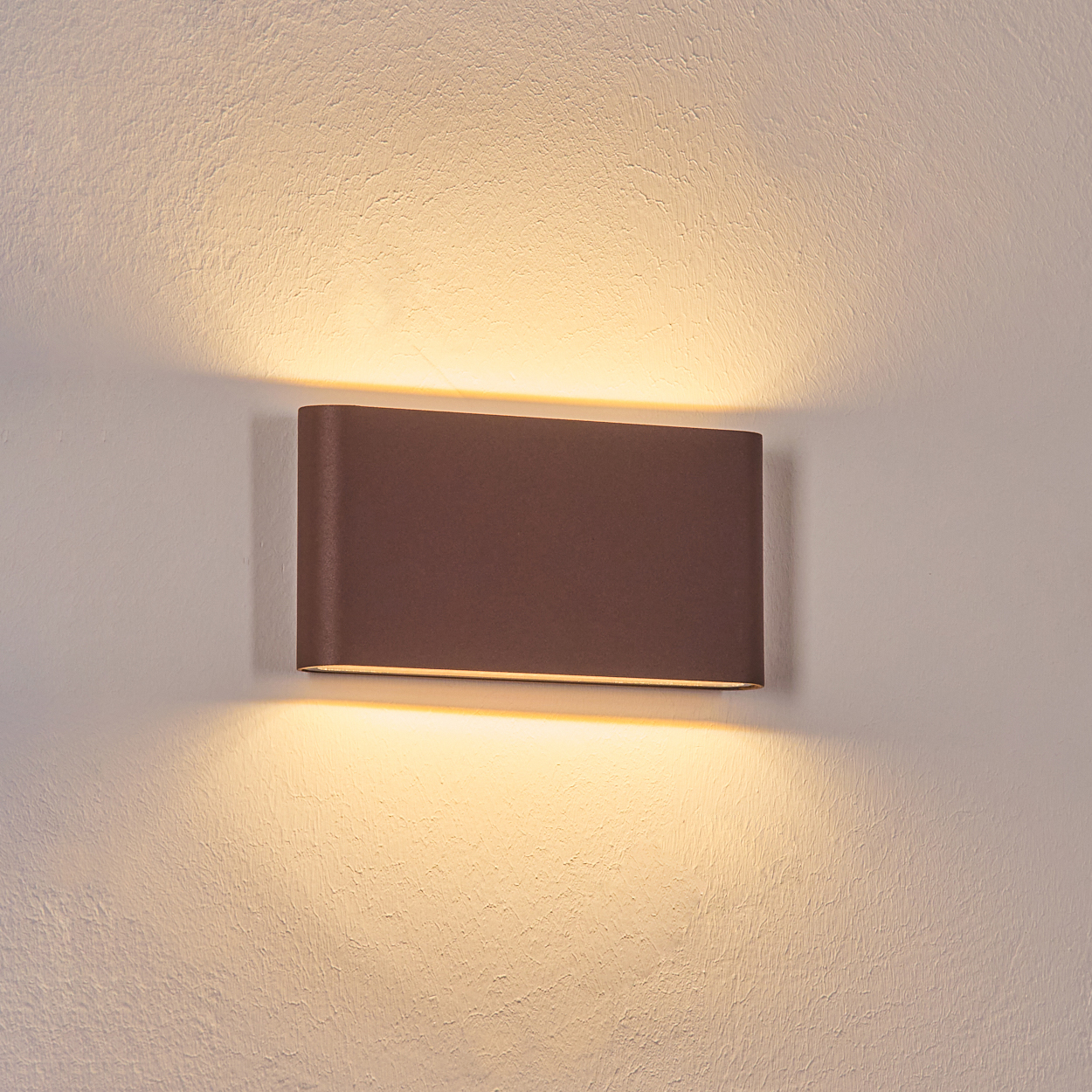 Eine braune, rechteckige Wandleuchte ist an einer strukturierten weißen Wand montiert und beleuchtet die Wand mit warmem, weichem Licht.