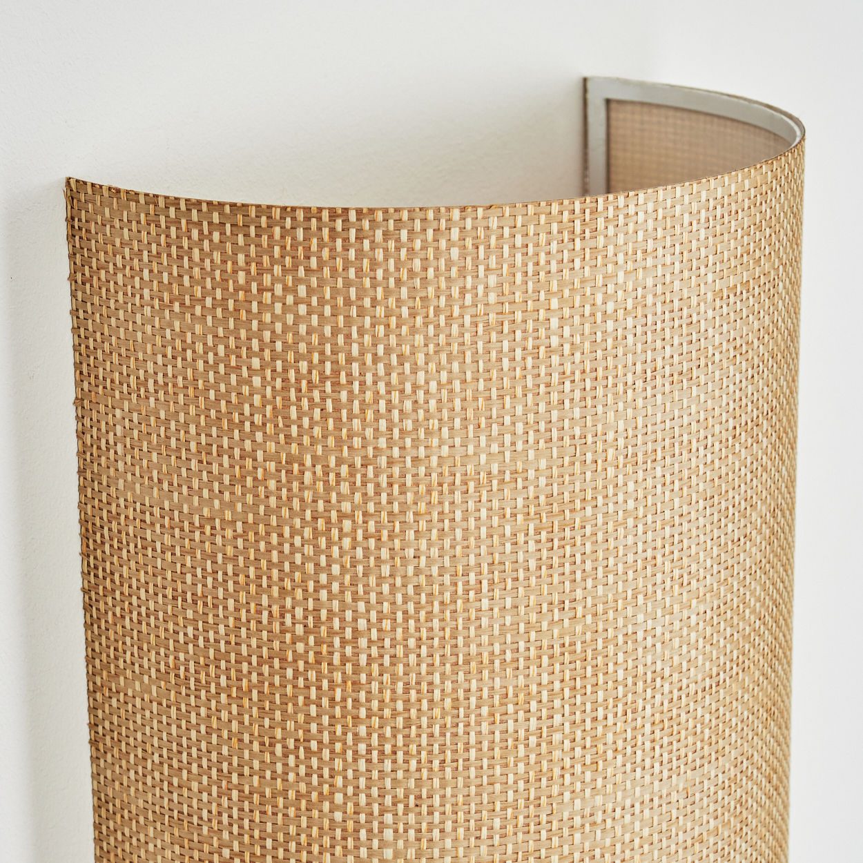 Eine Wandlampe aus gewebtem Rattan ist an einer weißen Wand befestigt. Die Lampe hat eine gebogene Form. Der Metallrahmen ist oben sichtbar.