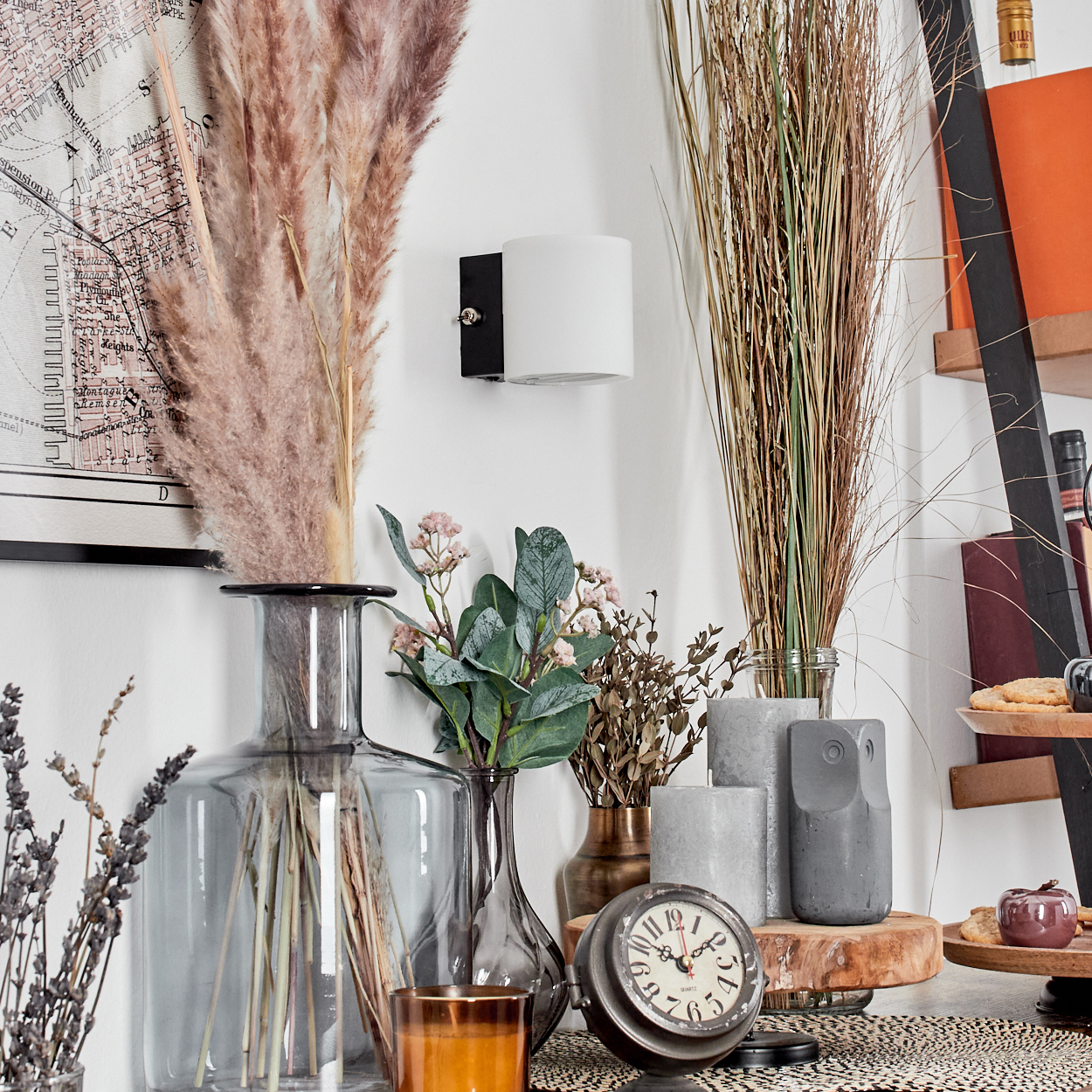 Eine dekorative Ausstellung mit Vasen, getrockneten Pflanzen, einer Uhr, Kerzen und einer Wandlampe. Im Hintergrund ist eine Karte zu sehen.