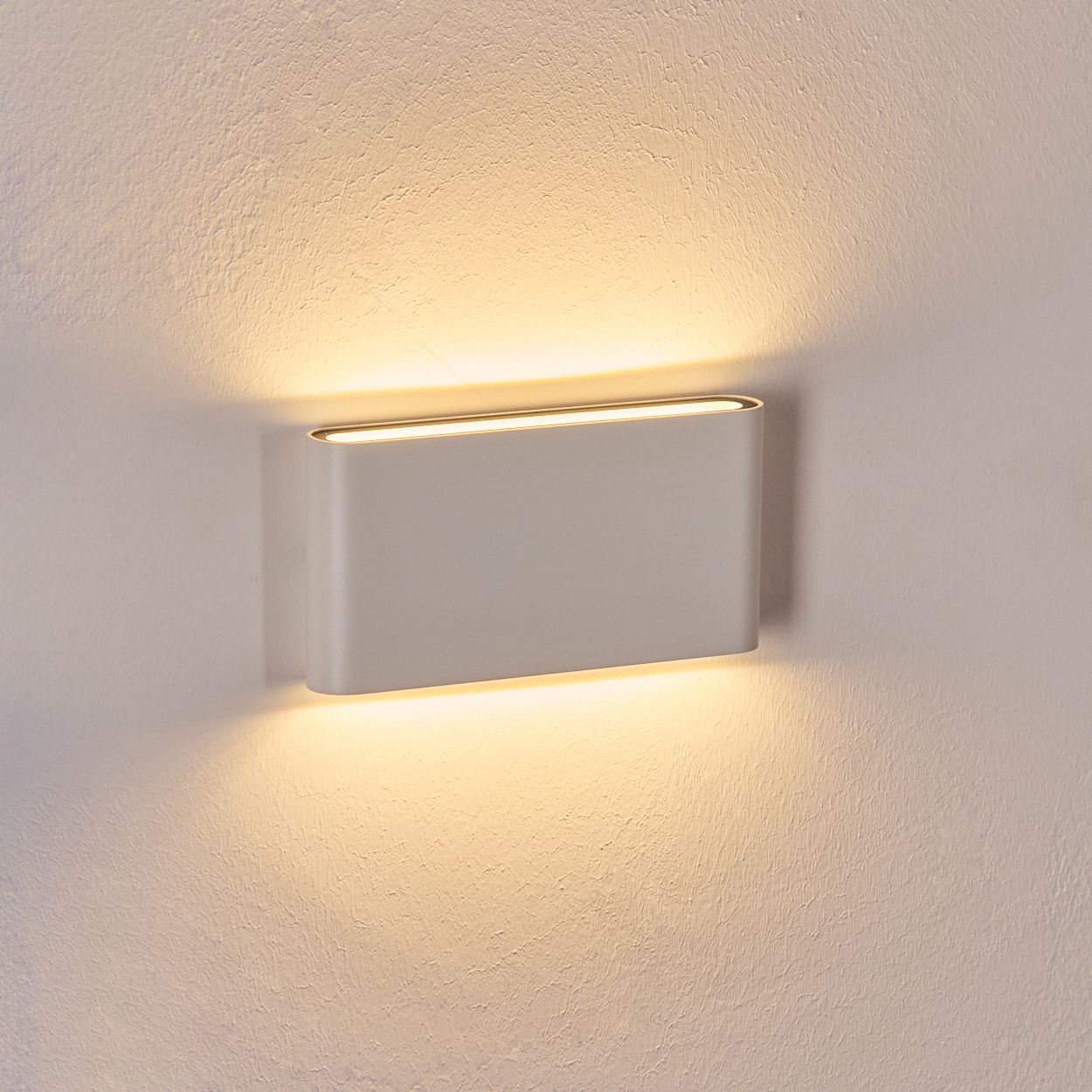 Eine rechteckige, weiße Wandleuchte, die warmes Licht ausstrahlt. Sie ist an einer strukturierten, cremefarbenen Wand montiert.