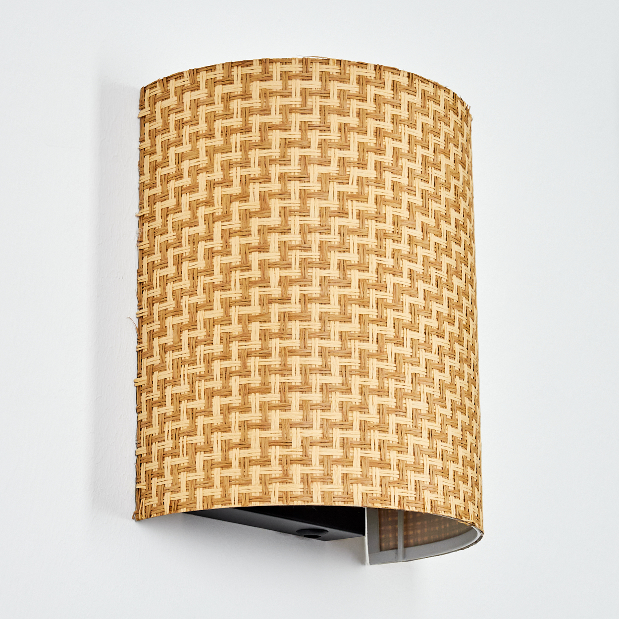 Eine gewebte, zylindrische Wandleuchte ist an einer weißen Wand befestigt. Die Lampe hat einen schwarzen Sockel und einen hellbraunen, gewebten Schirm.