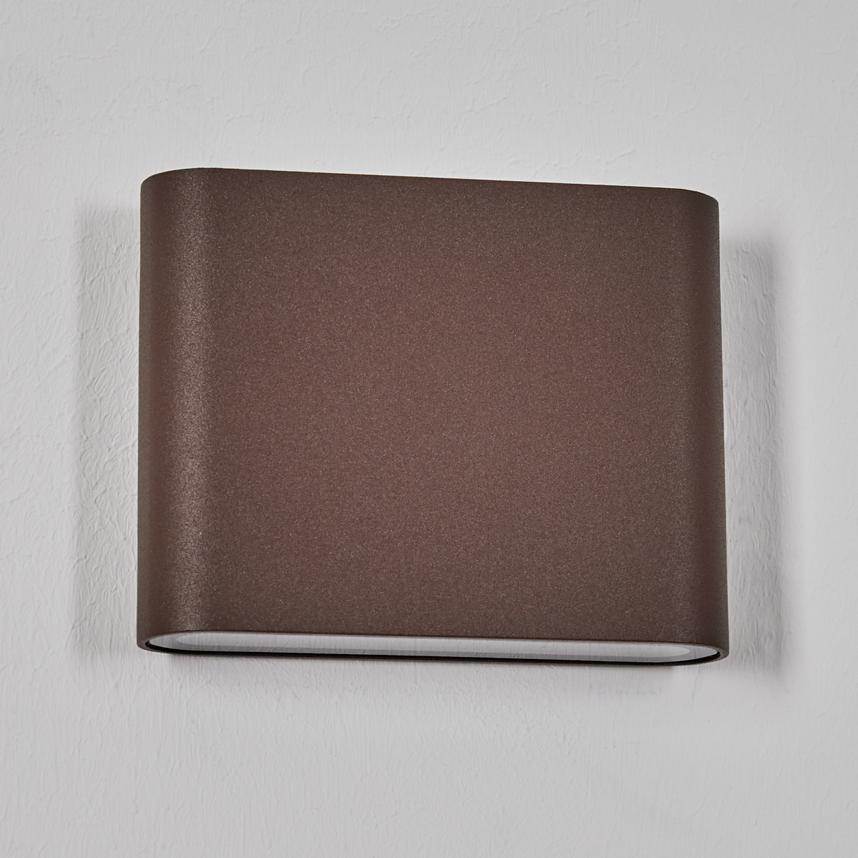 Eine braune, rechteckige Wandleuchte ist an einer weißen, strukturierten Wand befestigt. Die Lampe hat eine abgerundete Form und eine Öffnung.