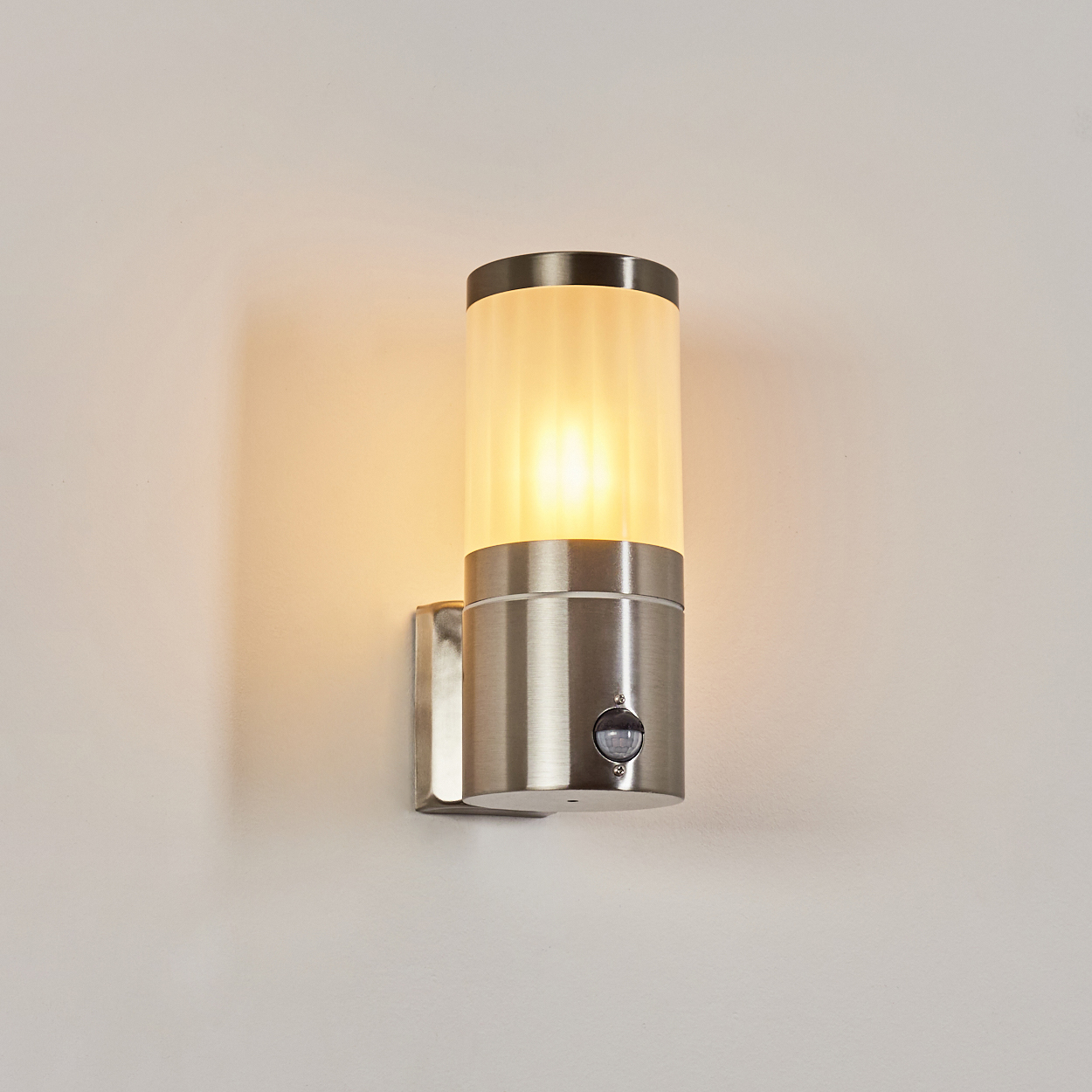 Eine wandmontierte, zylindrische Leuchte mit mattiertem Glasschirm und Metallsockel, die die Wand beleuchtet.