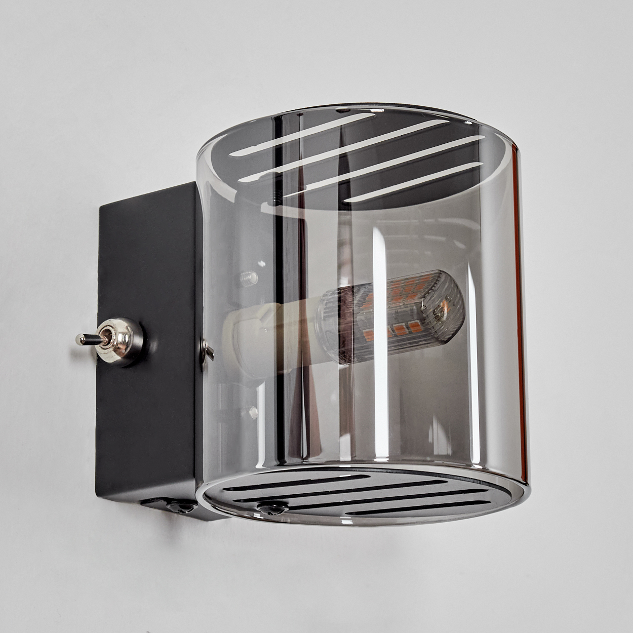 Eine moderne Wandleuchte mit zylindrischem Glasschirm, schwarzem Metallsockel und Chromschalter, an einer weißen Wand montiert.