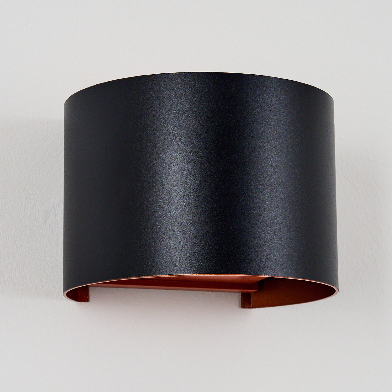 Eine moderne schwarze Wandlampe mit kupferfarbenem Innenraum ist an einer weißen Wand montiert. Die Lampe hat eine gebogene Form.