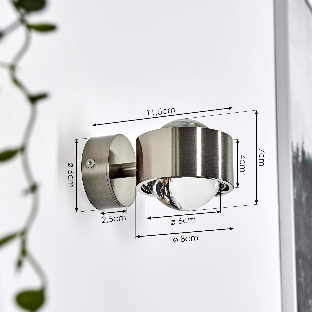 Eine metallische Wandleuchte ist an einer weißen Wand montiert. Abmessungen sind mit Pfeilen angegeben. Grüne Pflanze links, ein Gemälde rechts.