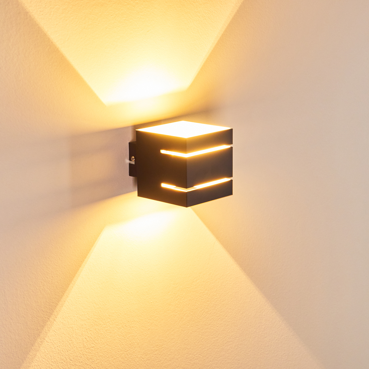 Eine moderne schwarze Wandleuchte, die warmes Licht auf eine strukturierte weiße Wand wirft. Das Licht erzeugt helle, eckige Muster.