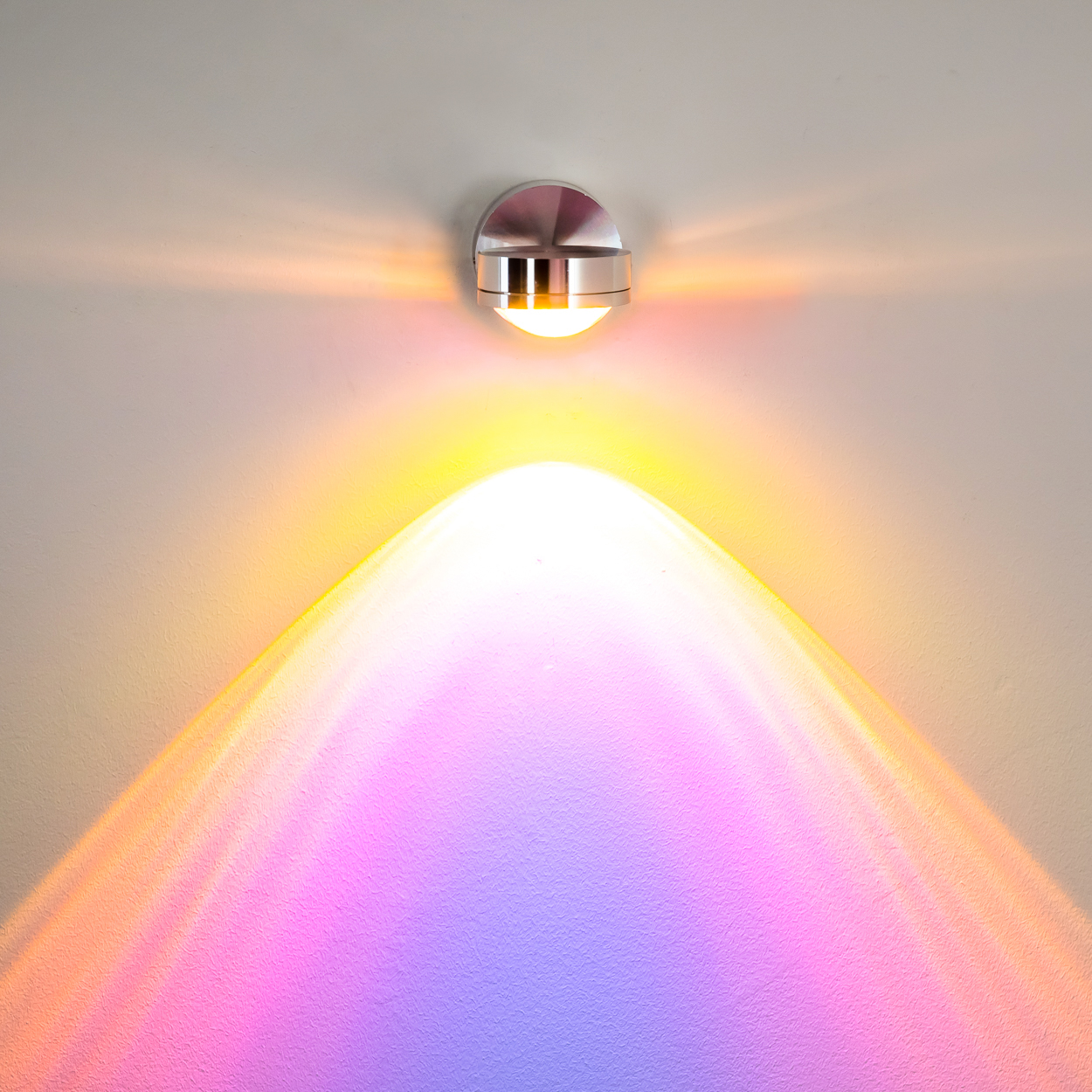 Eine moderne Wandlampe strahlt farbenfrohes Licht auf eine strukturierte Wand. Das Licht erzeugt einen Farbverlauf von Rosa nach Gelb.