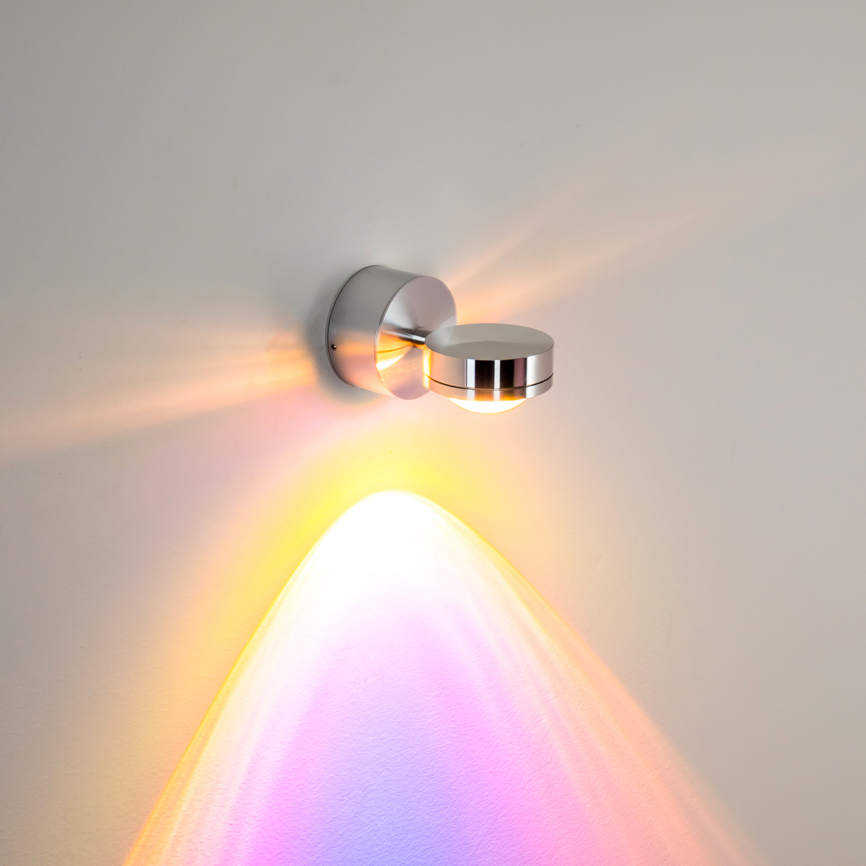 Eine moderne Metall-Wandleuchte wirft farbiges Licht auf eine weiße Wand. Das Licht erzeugt ein lebendiges, bogenförmiges Muster.