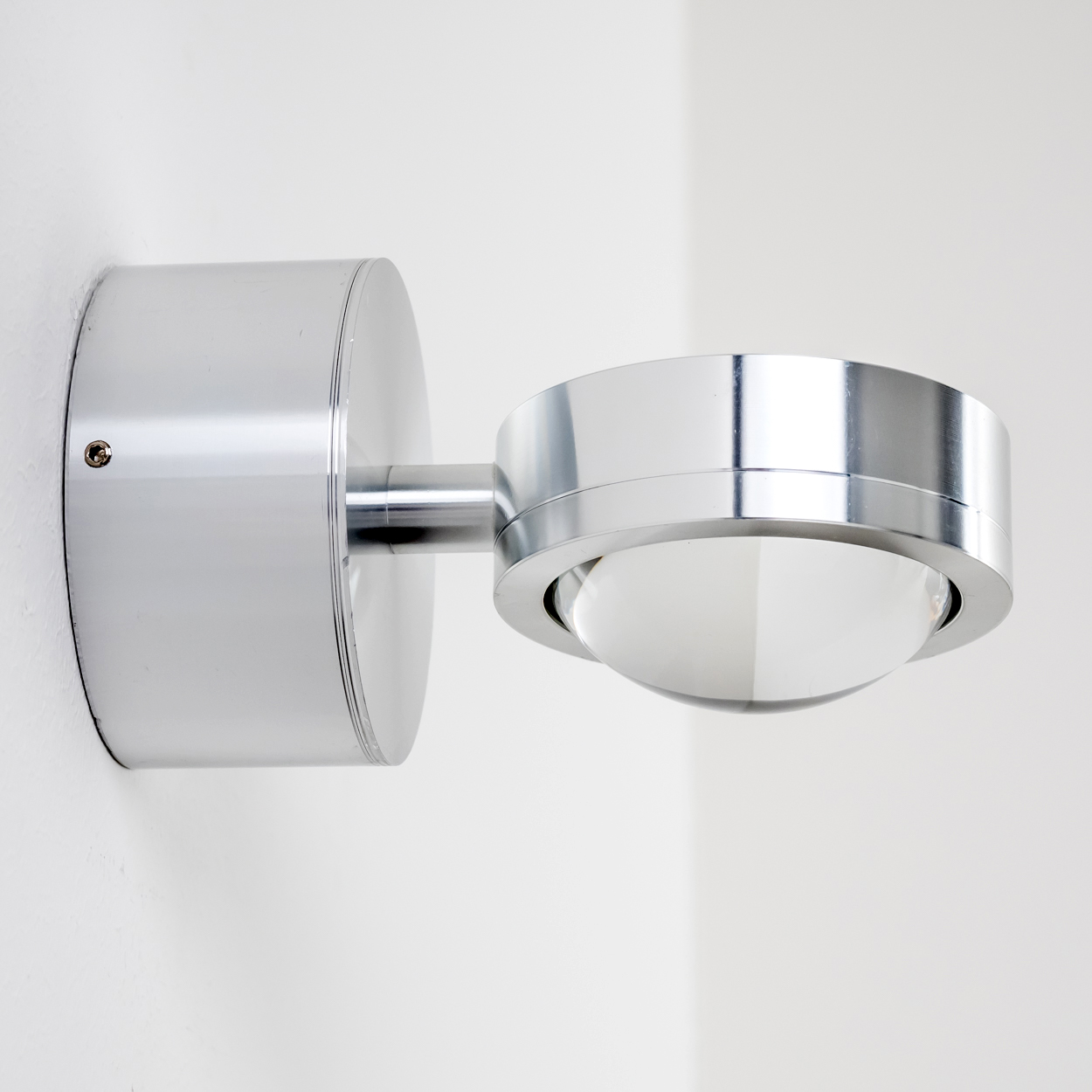 Eine moderne, silberne Wandleuchte ist an einer weißen Wand befestigt. Die Lampe hat einen runden Sockel, einen Verbindungsarm und ein kugelförmiges Licht.