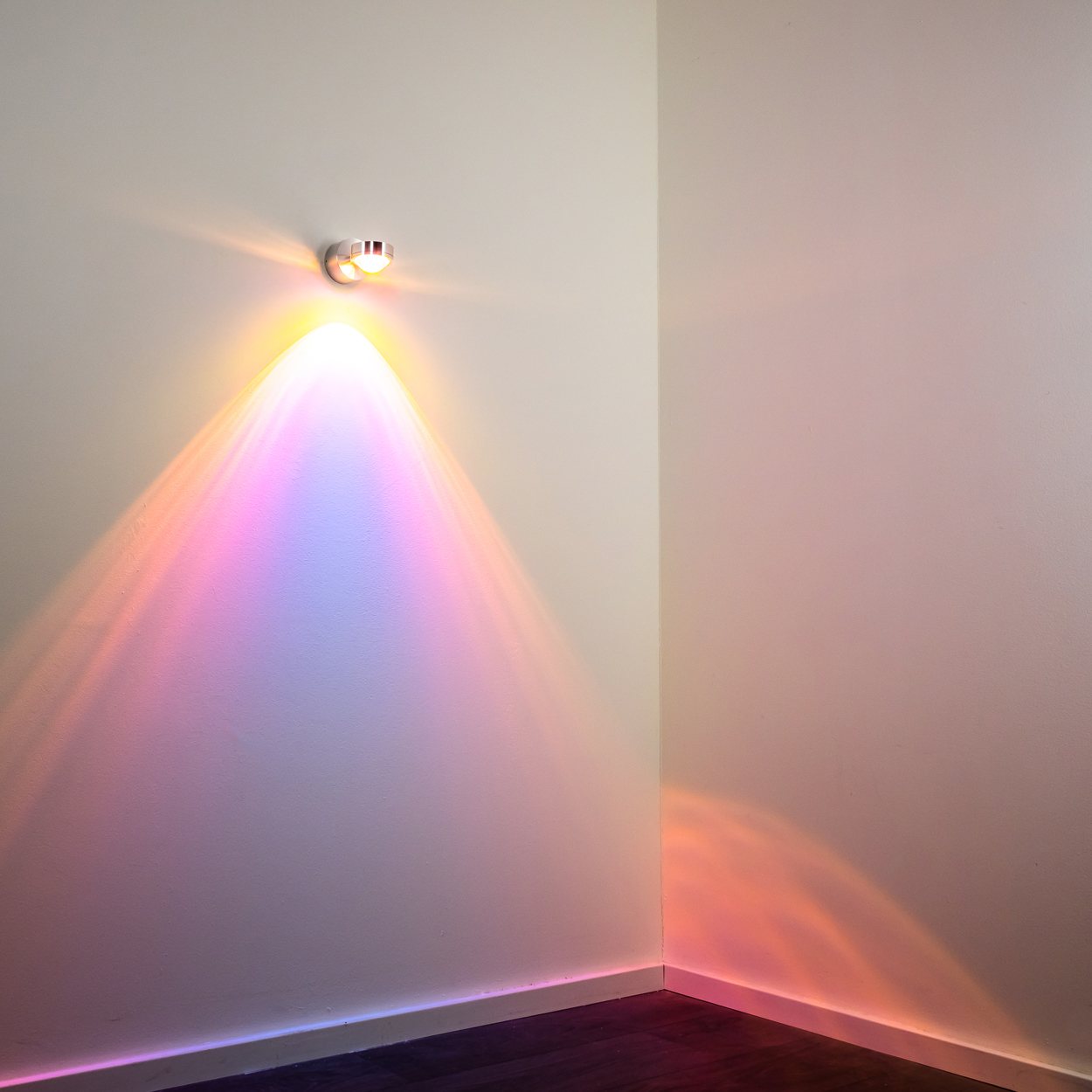Eine Wandleuchte projiziert einen farbenfrohen Bogen in die Ecke eines Raumes und erzeugt ein Spektrum aus Rosa, Lila und Orangetönen.