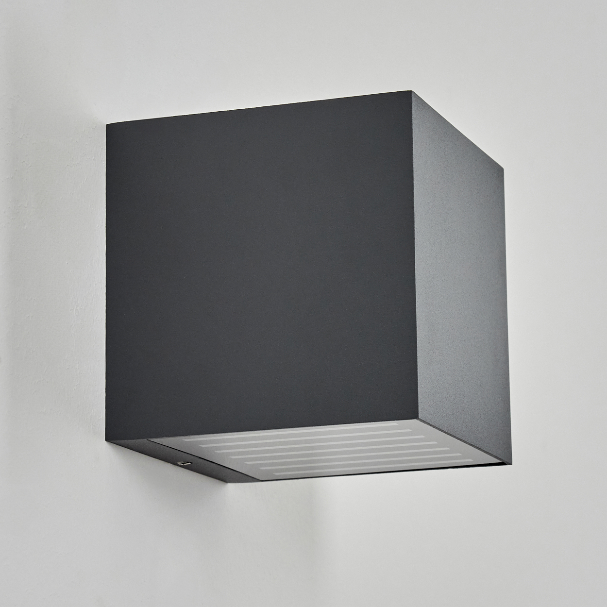 Eine dunkelgraue, würfelförmige Wandleuchte ist an einer weißen Wand befestigt. Die Lampe hat eine Lichtquelle an der Unterseite.