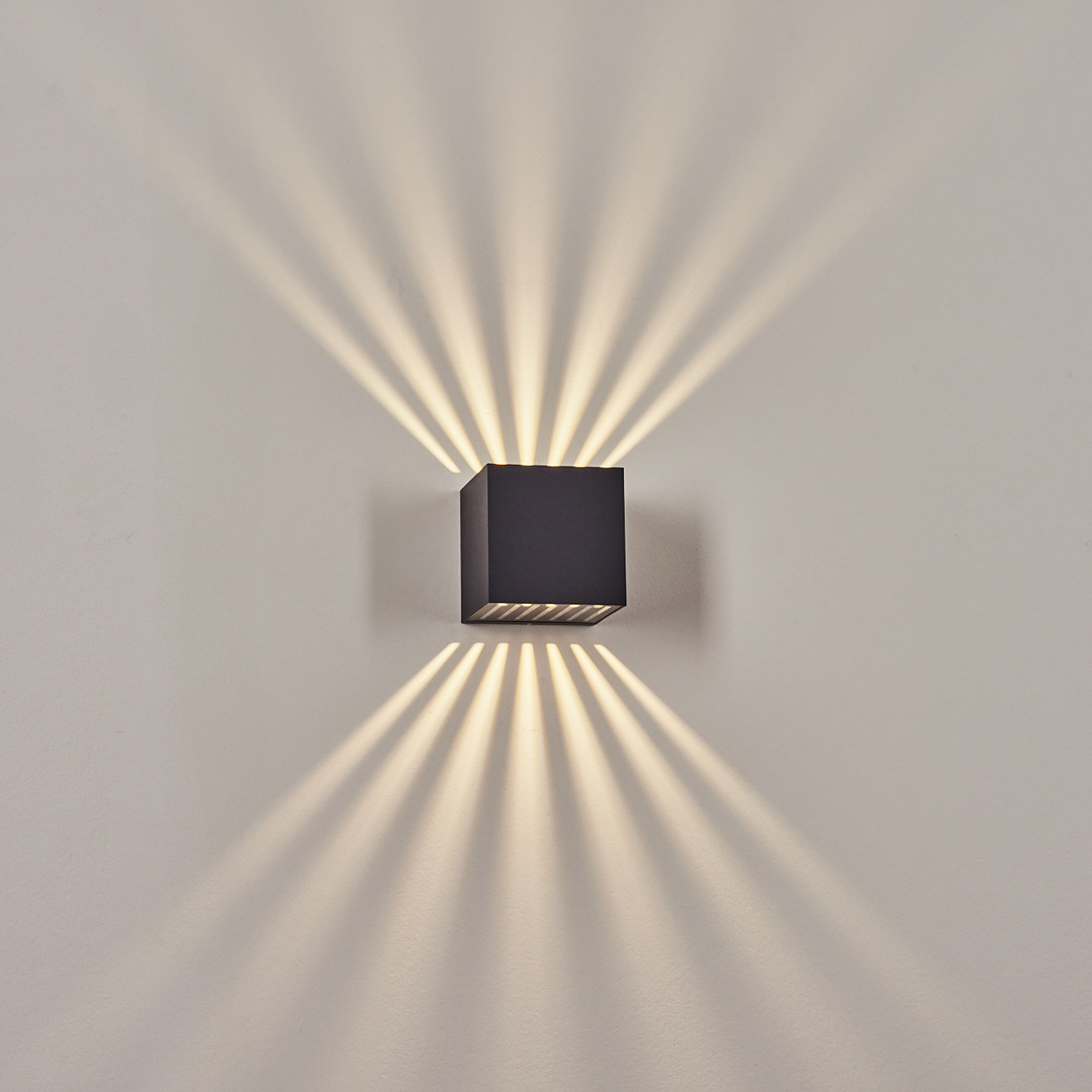Eine quadratische, schwarze Wandleuchte strahlt Licht in mehreren Strahlen aus. Das Licht erzeugt ein fächerartiges Muster an der beigen Wand.