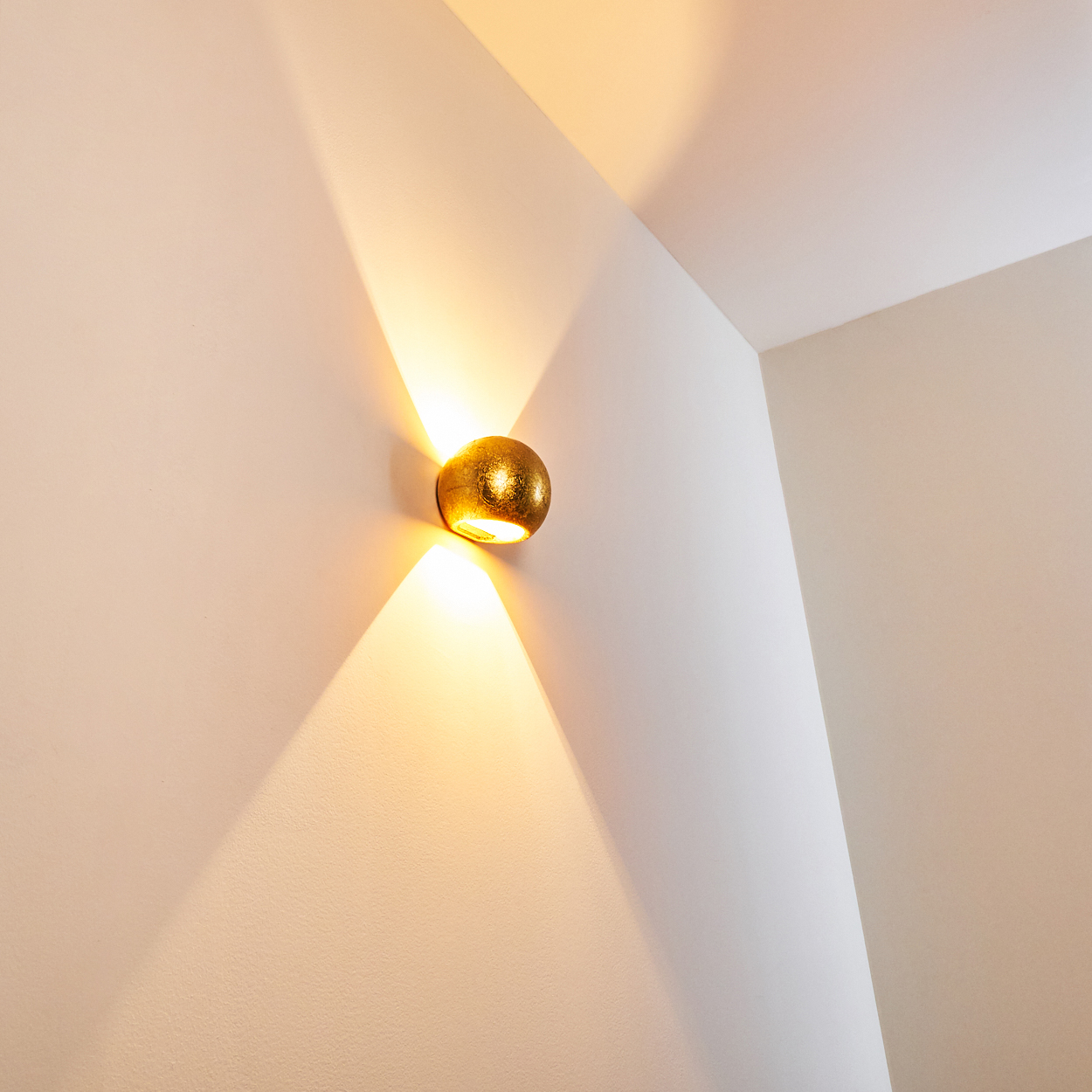 Eine goldene, strukturierte Wandleuchte strahlt nach oben und unten und wirft Schatten auf die weißen Wände einer Ecke.