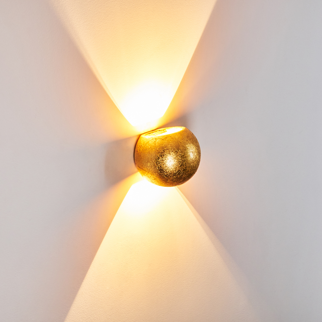 Eine goldene, kugelförmige Wandleuchte ist an einer weißen Wand befestigt. Die Lampe strahlt zwei Lichtstrahlen aus, die die Wand beleuchten.