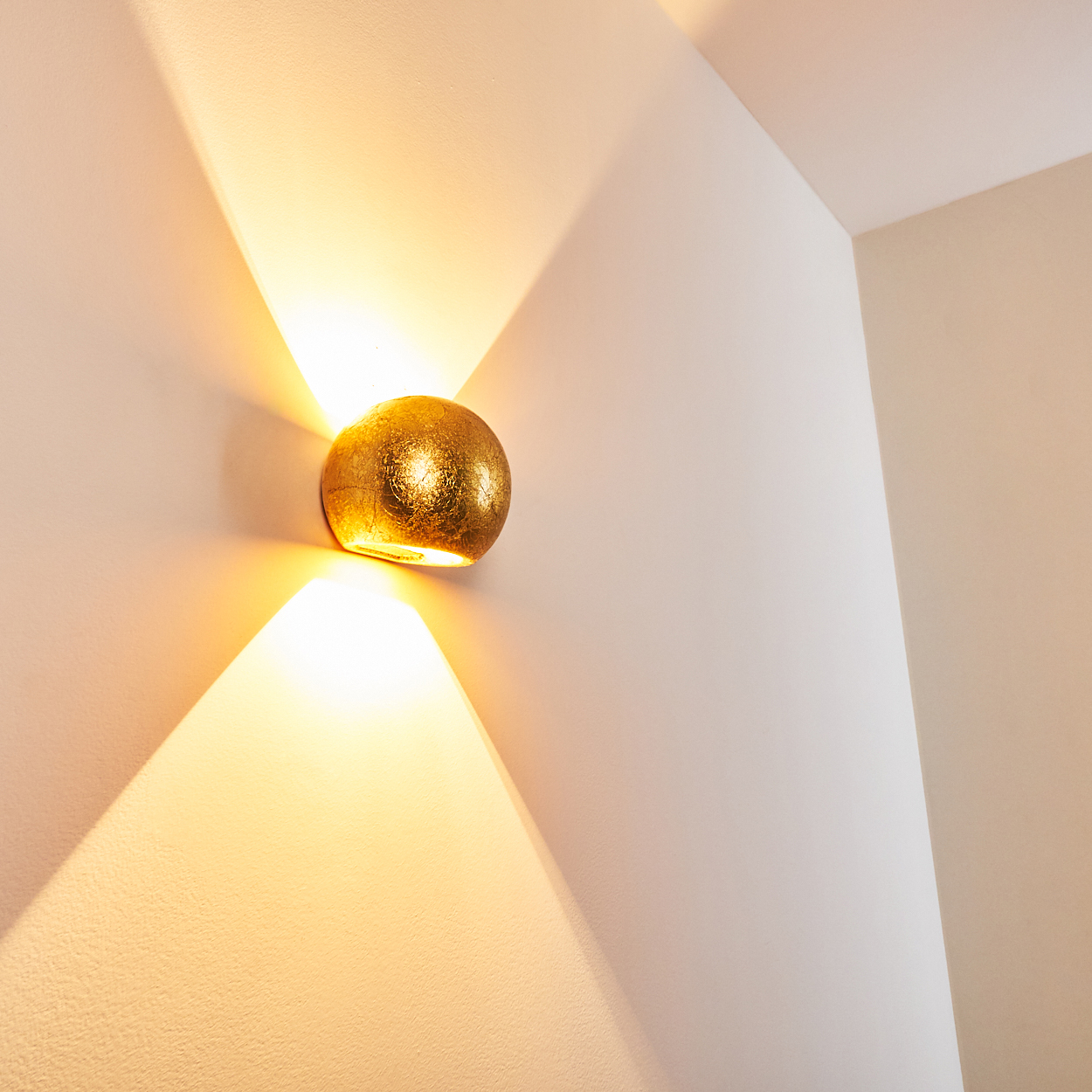 Eine goldene, strukturierte Wandleuchte ist an einer weißen Wand befestigt. Die Lampe wirft Licht nach oben und unten und erzeugt ein dreieckiges Leuchten.