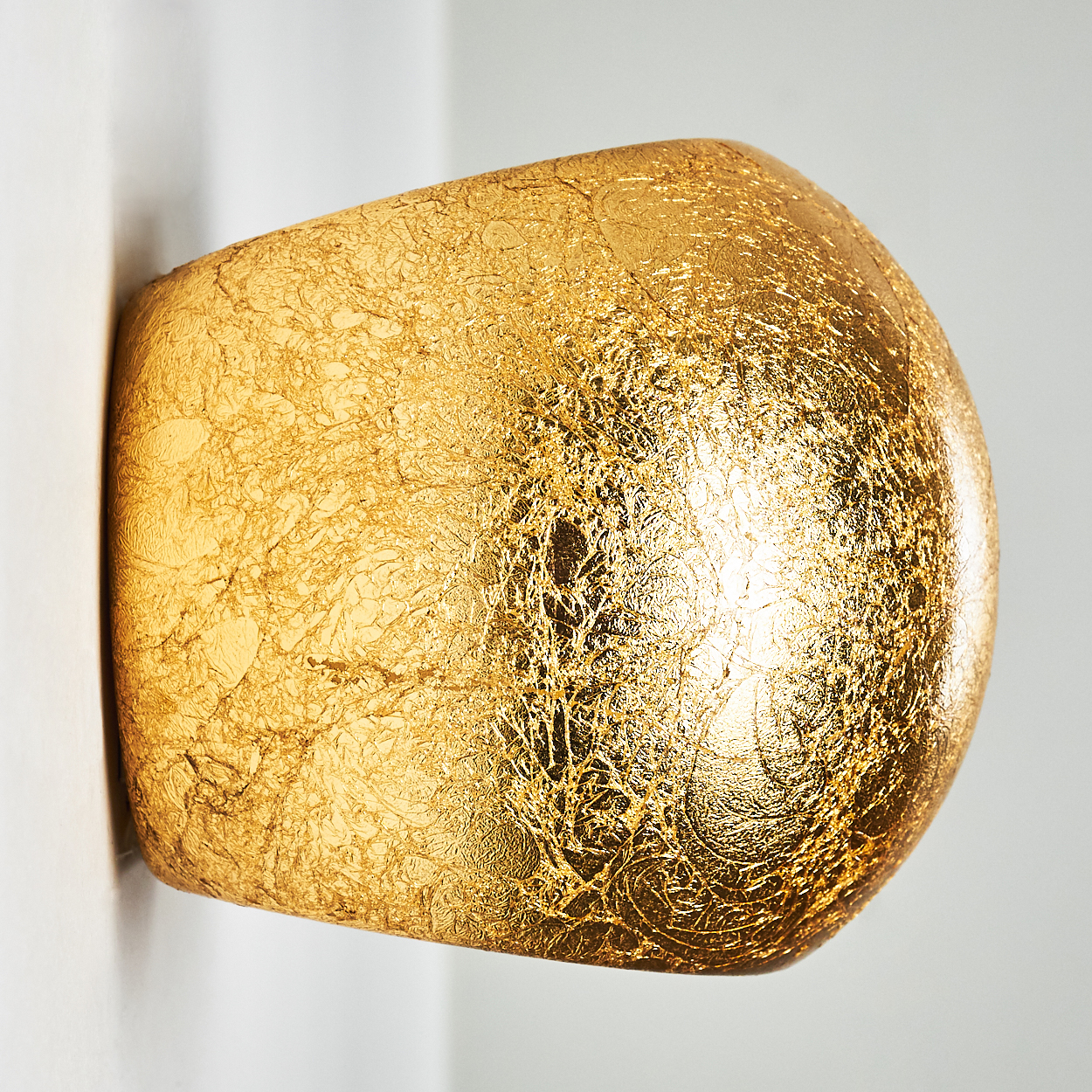 Eine goldene, strukturierte Wandleuchte ist an einer weißen Wand befestigt. Die Leuchte hat eine abgerundete, unregelmäßige Form.