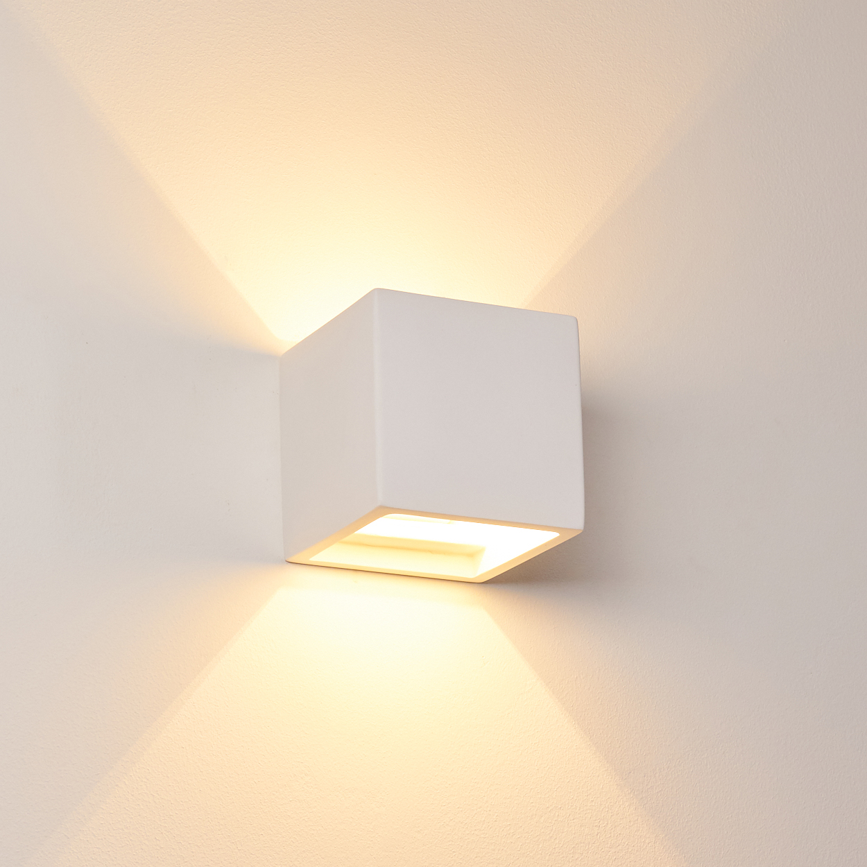 Eine weiße quadratische Wandleuchte strahlt warmes Licht aus. Das Licht breitet sich nach oben und unten an der Wand aus. Die Wand ist weiß.