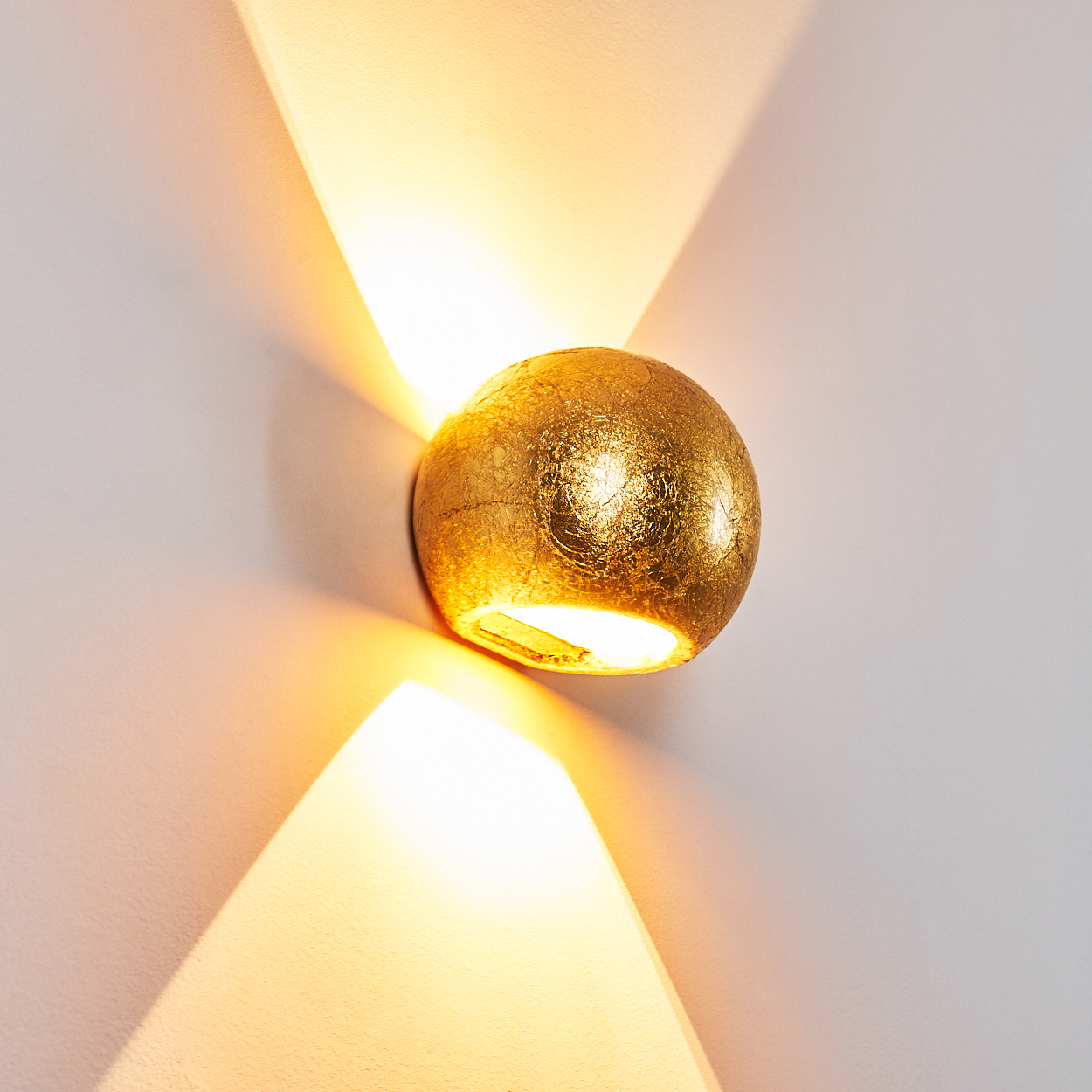 Eine goldene, kugelförmige Wandleuchte ist an einer weißen Wand befestigt. Licht strahlt nach außen und erzeugt ein sternförmiges Muster.