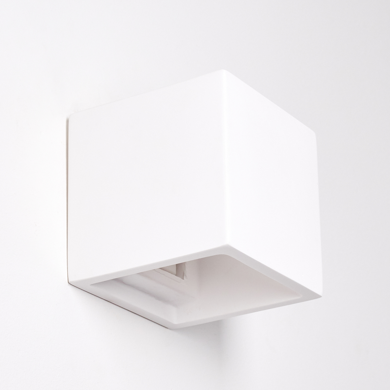 Eine weiße, kubusförmige Wandleuchte ist an einer weißen Wand befestigt. Die Lampe hat eine offene Unterseite, die ihr Inneres zeigt.