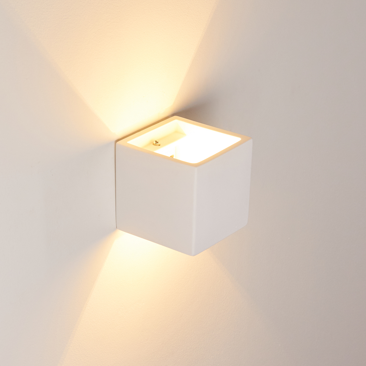 Eine weiße, würfelförmige Wandleuchte ist an einer weißen Wand montiert. Sie strahlt helles Licht nach oben und unten aus.