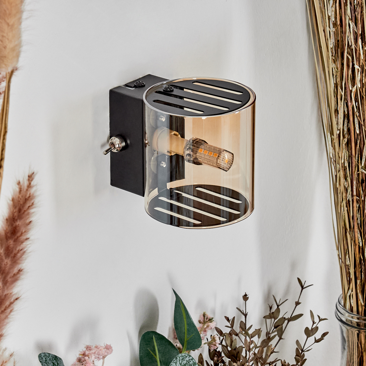 Eine an der Wand montierte schwarz-goldene Leuchte mit Glühbirne, umgeben von dekorativen Pflanzen. Ein Kippschalter ist sichtbar.