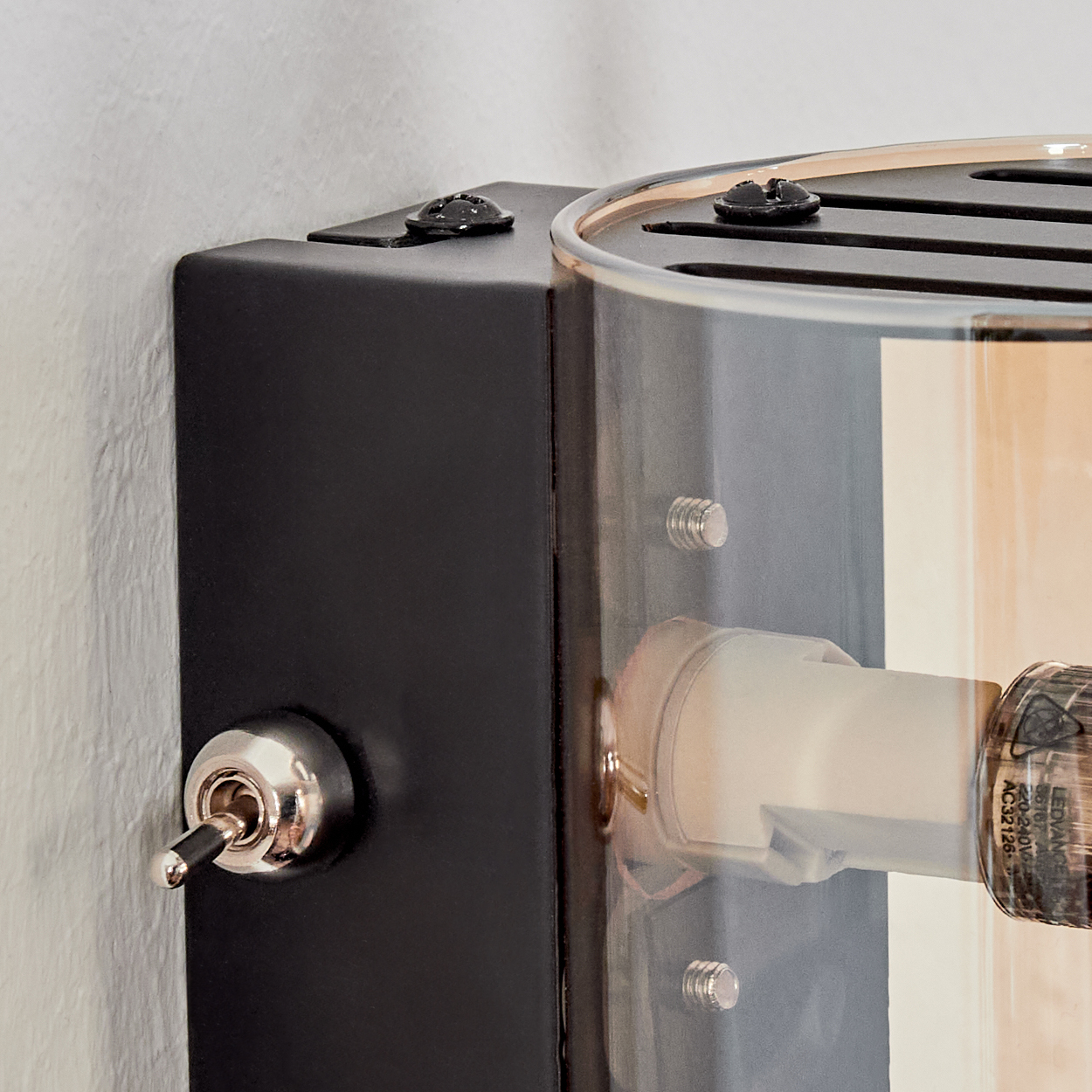 Nahaufnahme einer schwarzen Wandleuchte mit Kippschalter, transparentem Glas und einer Glühbirne. Die Lampe ist an einer weißen Wand befestigt.