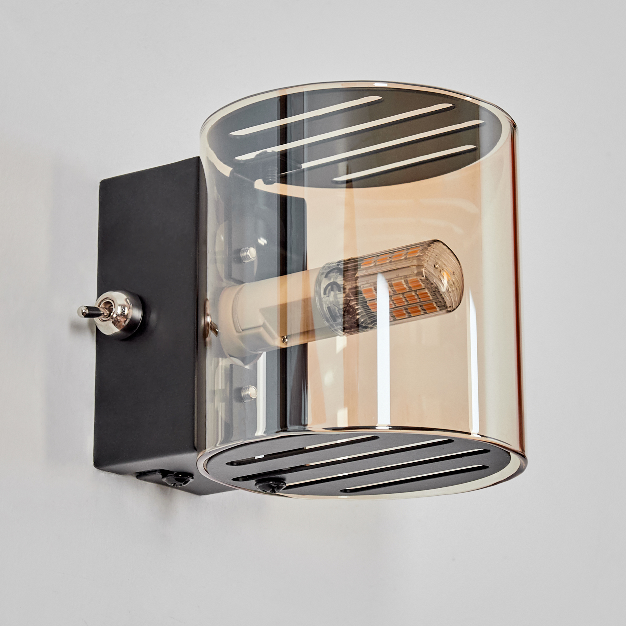 Eine moderne Wandleuchte mit schwarzem Sockel, einem Kippschalter und einem zylindrischen, bernsteinfarben getönten Glasschirm mit sichtbarer Glühbirne.
