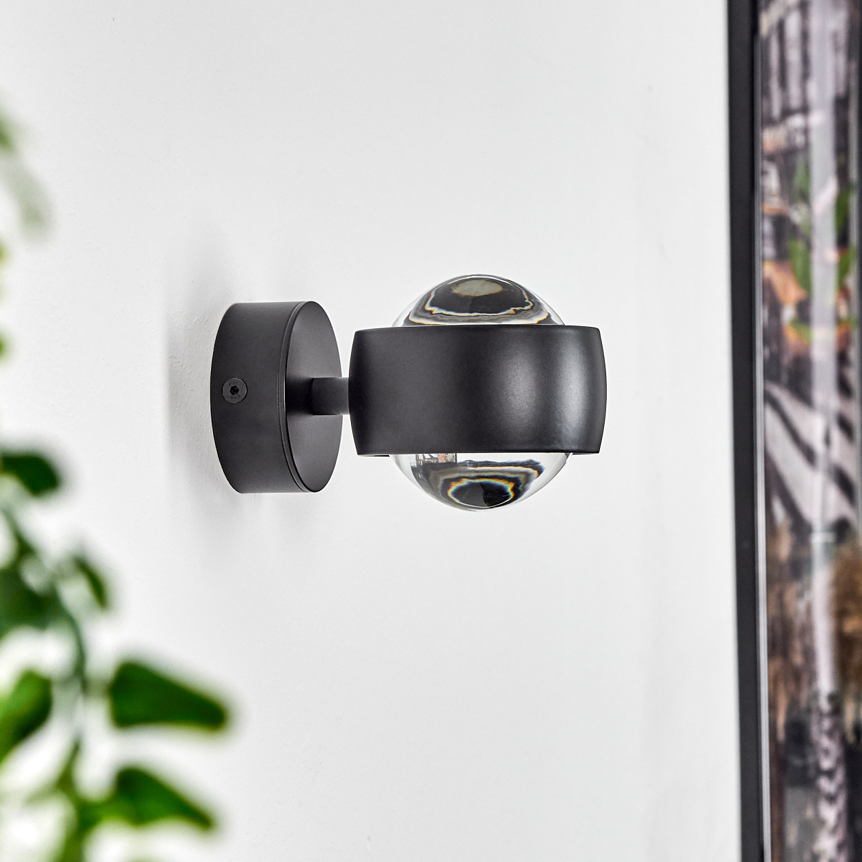 Eine moderne schwarze Wandleuchte mit kugelförmigem Glasschirm ist an einer weißen Wand befestigt. Einige grüne Blätter sind im Vordergrund.